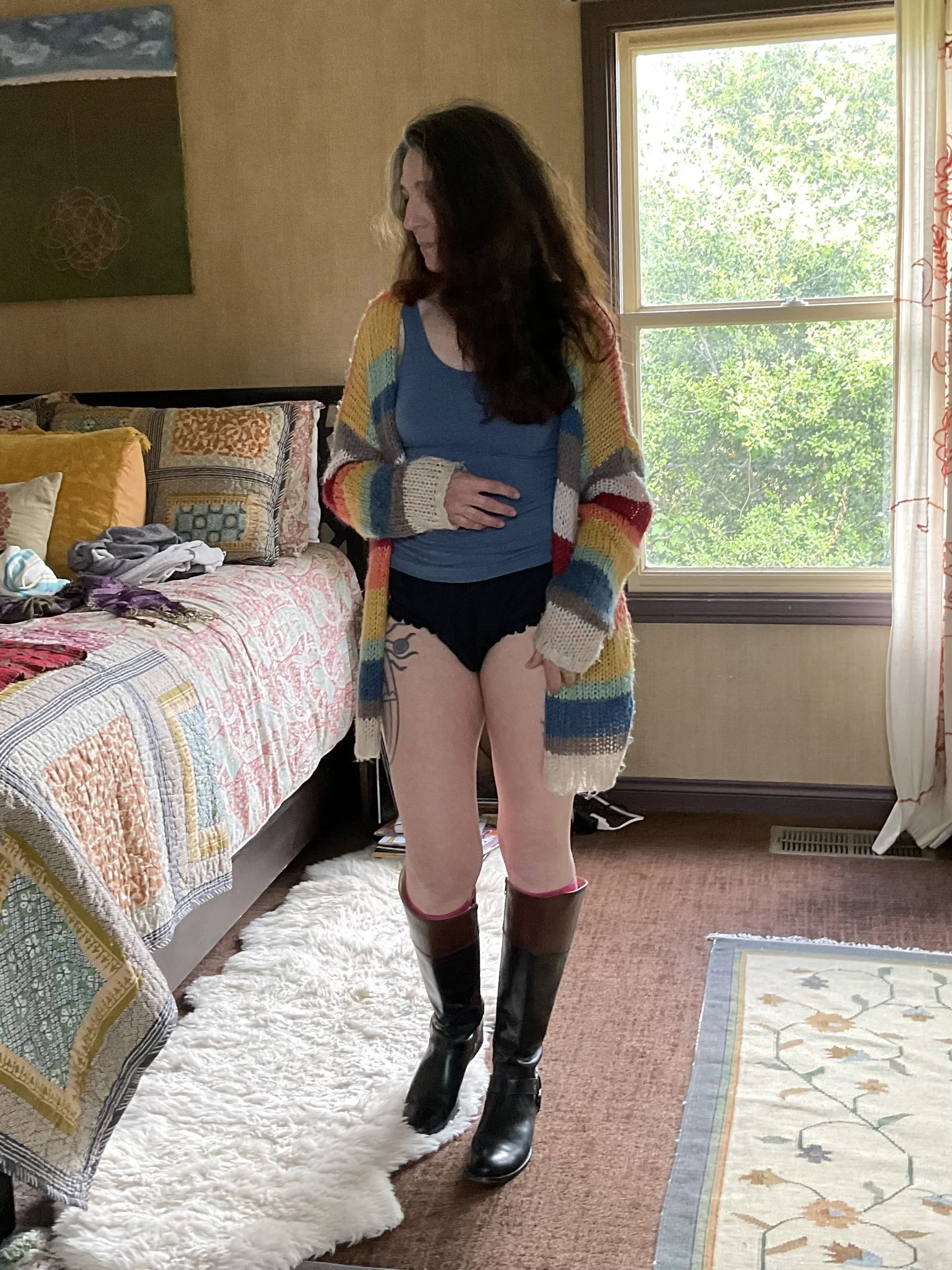 Woman in sustainable cheeky underwear, standing in a bedroom, and also wearing a striped cardigan and knee-high boots.