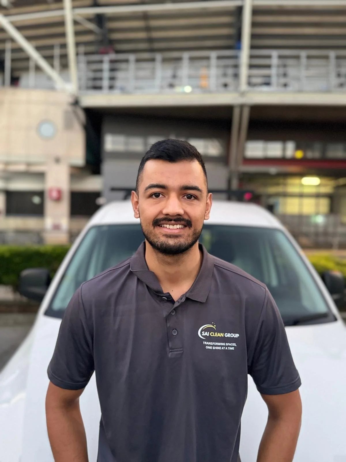 A man with short dark hair, beard, and mustache smiling at the camera, standing outdoors in front of a white vehicle, wearing a dark gray polo shirt with a logo that reads 'SAI CLEAN GROUP' and the slogan 'Transforming Spaces, One Shine at a Time.' Behind him, there is a building with multiple stories and a green shrubbery.