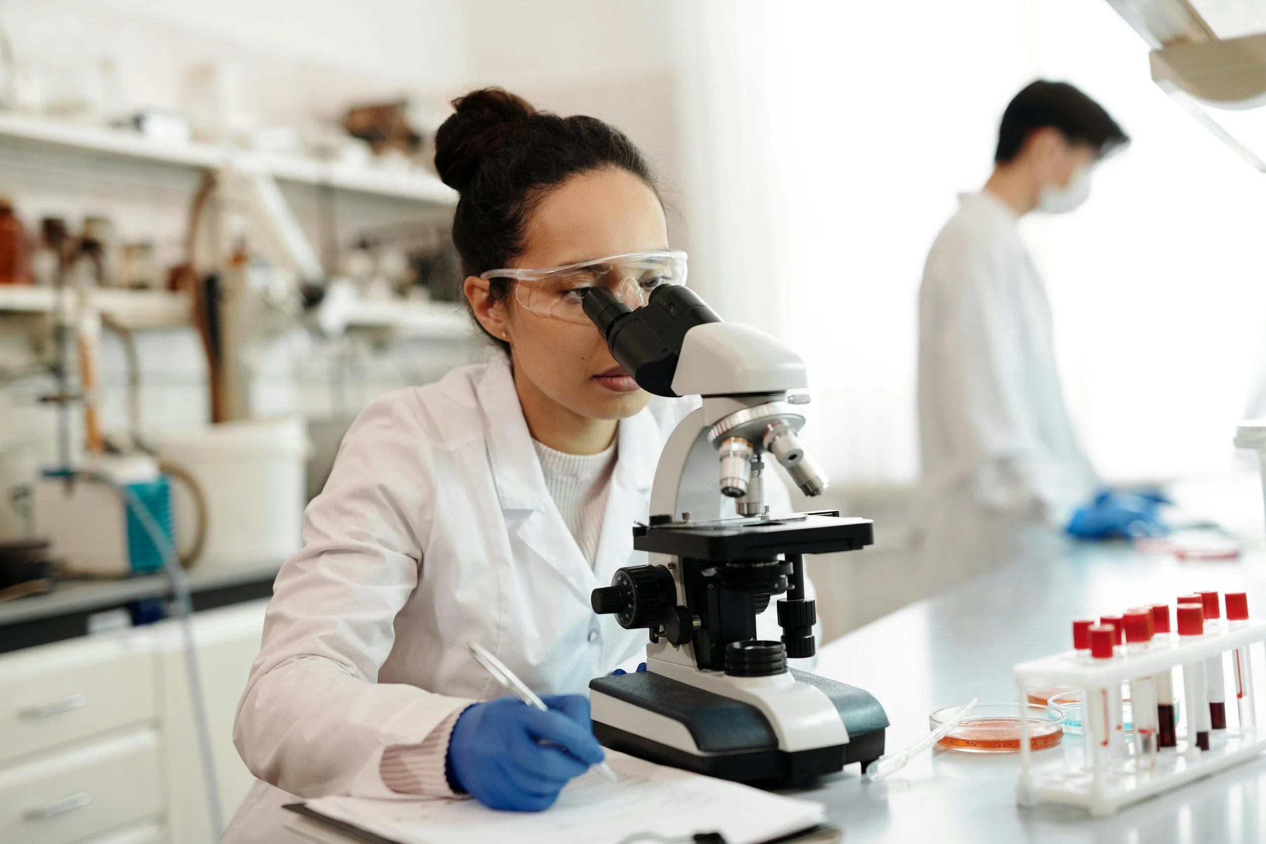 A female scientist in safety glasses and gloves looking through a microscope in a laboratory, with another scientist working in the background.