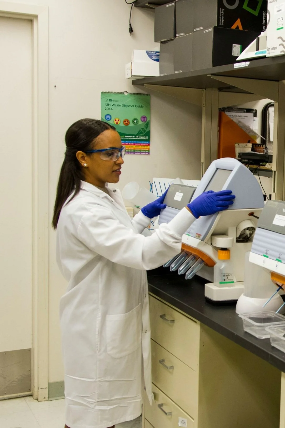 A woman scientist wearing safety goggles, gloves, and a lab coat operating a laboratory machine in a lab with shelves and a waste disposal guide on the wall.