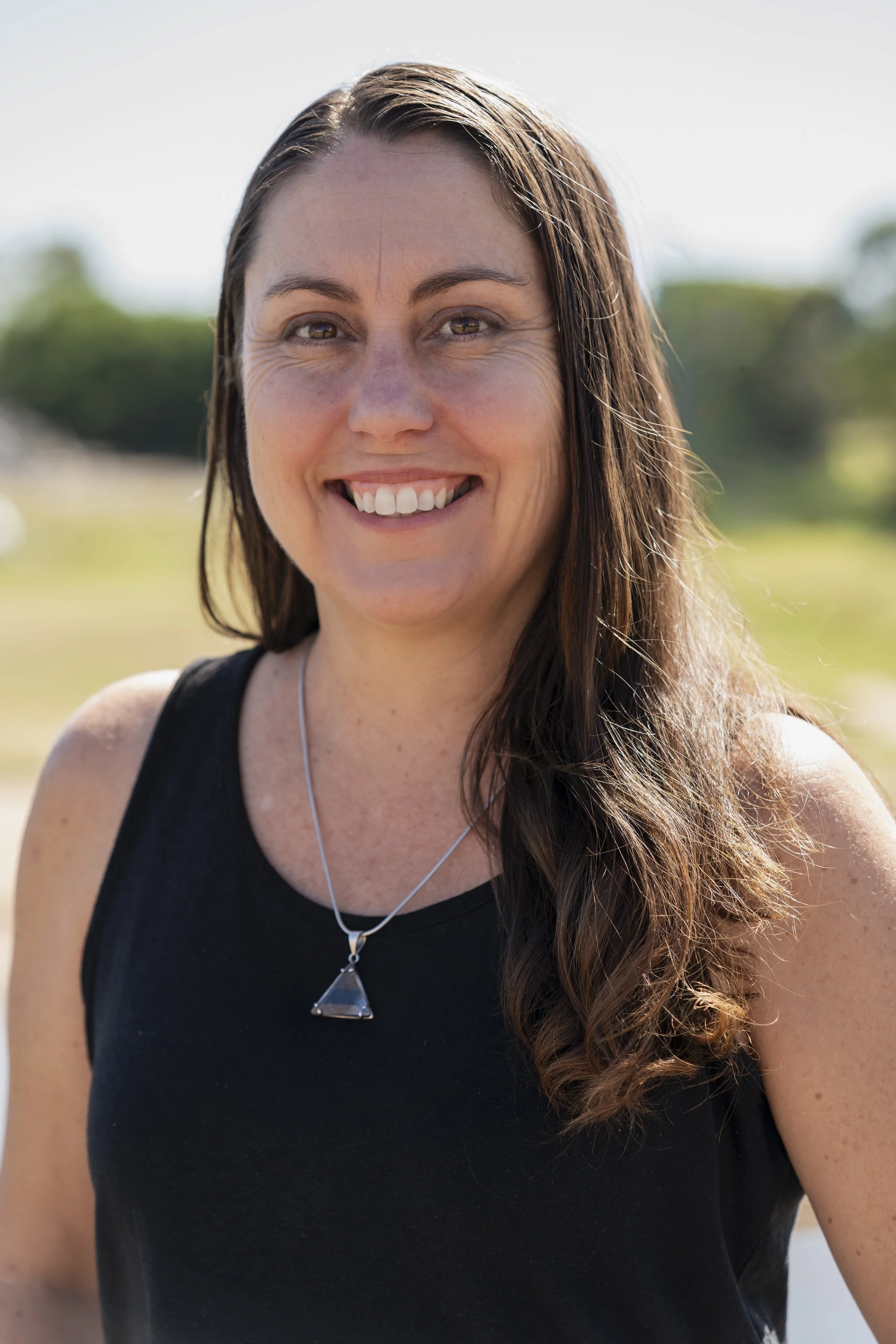 A smiling woman with long brown hair wearing a black sleeveless top and a silver necklace with a triangular pendant outdoors on a sunny day.