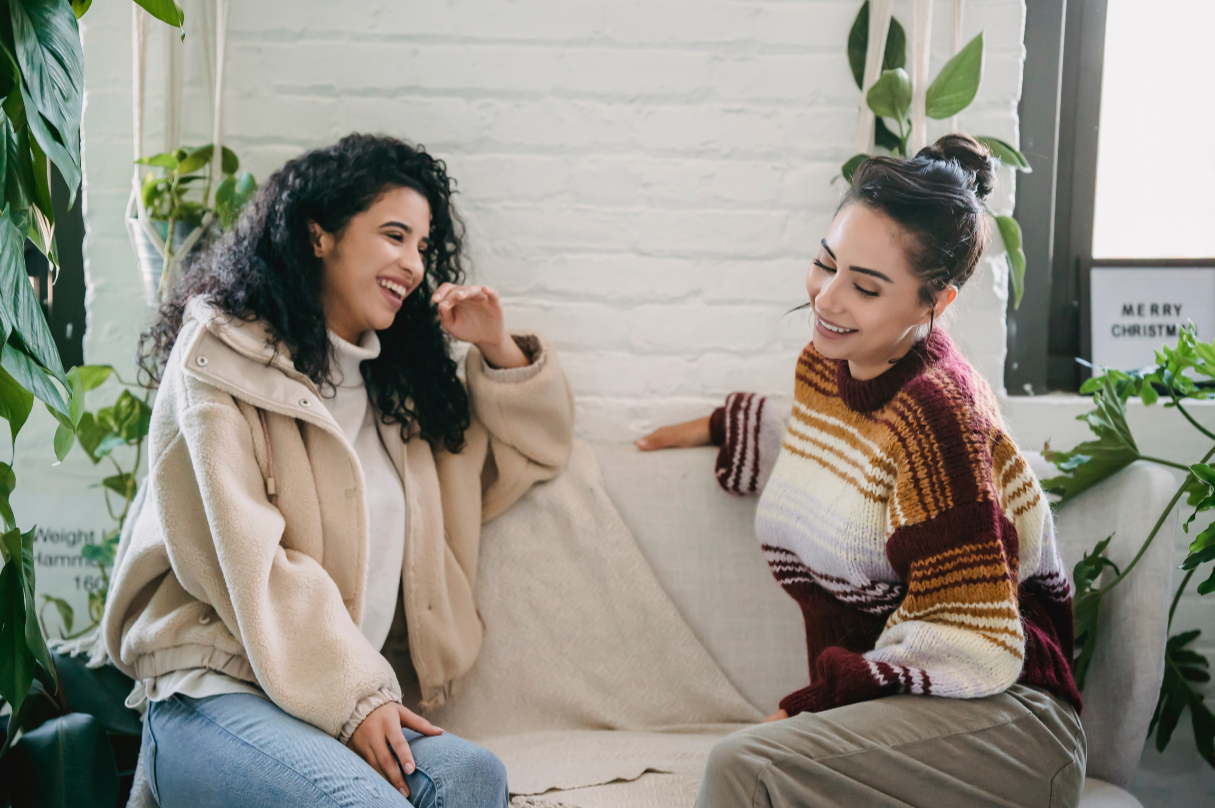 Two women sitting on a white couch, talking and smiling, surrounded by green plants in a cozy indoor setting.