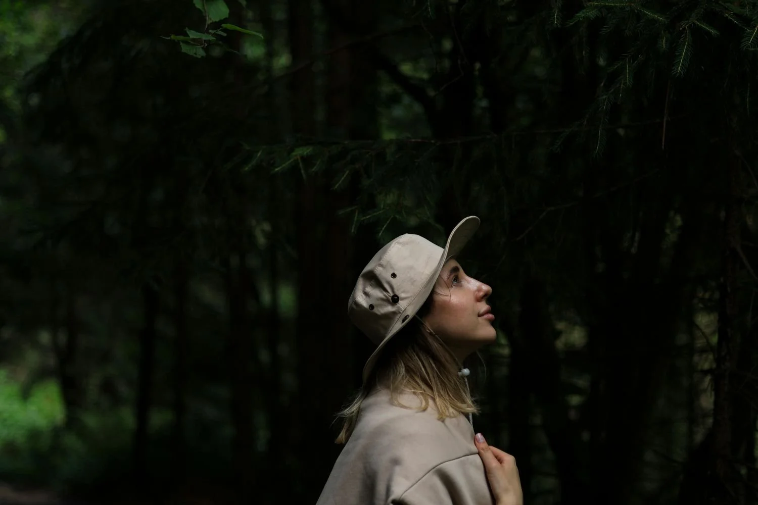 A woman in a beige hat and jacket standing in a dark forest, looking upward.