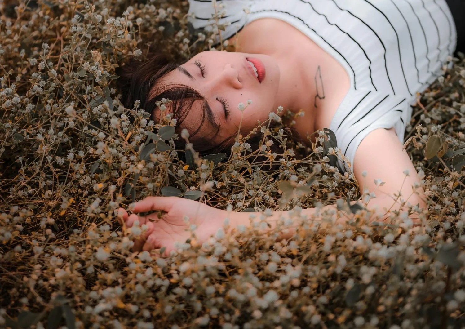A woman lying on her back on dry foliage, eyes closed, wearing a white and black striped shirt.