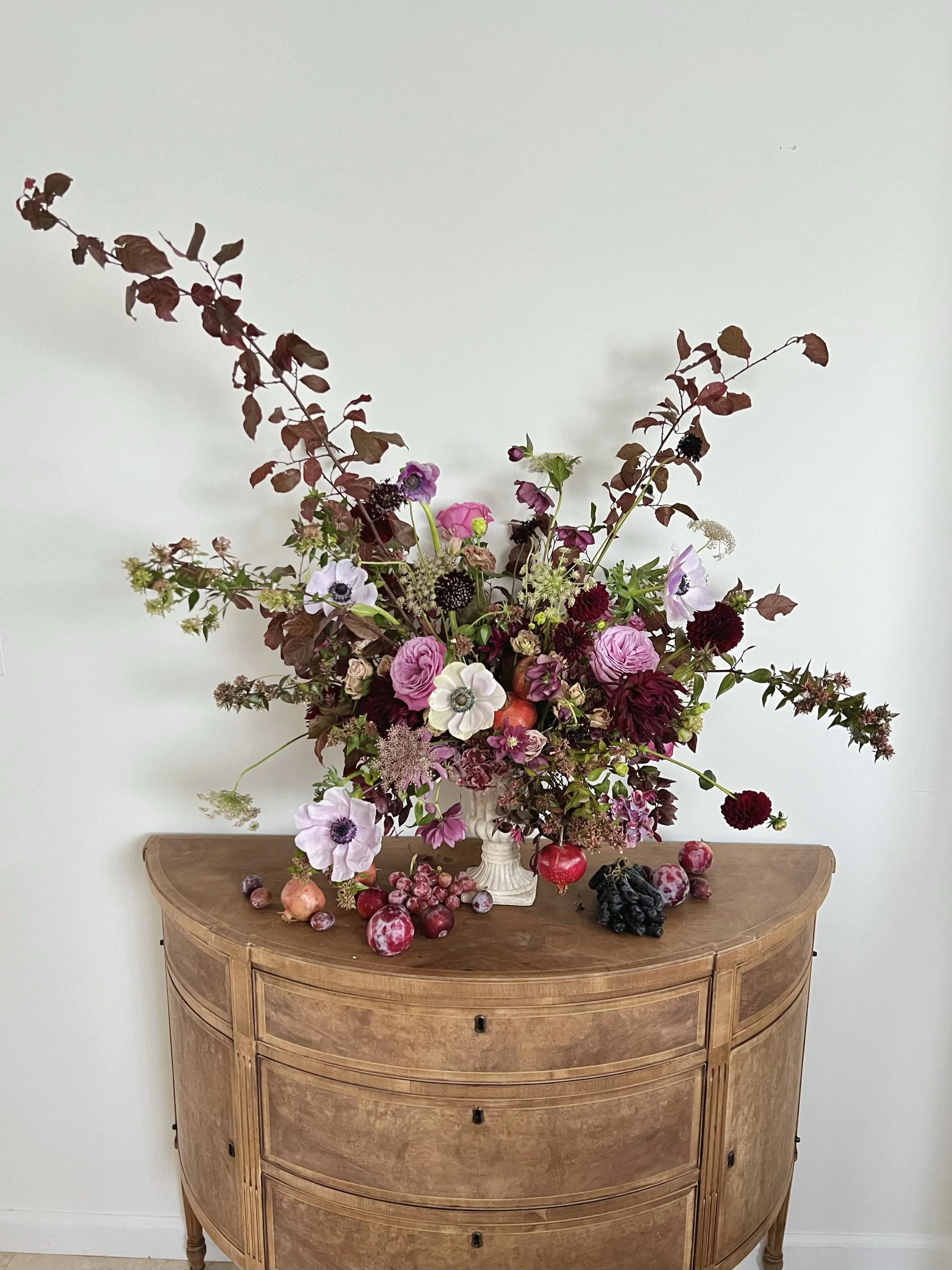 A wooden sideboard with a floral arrangement in a vase on top, surrounded by scattered grapes and pomegranates.