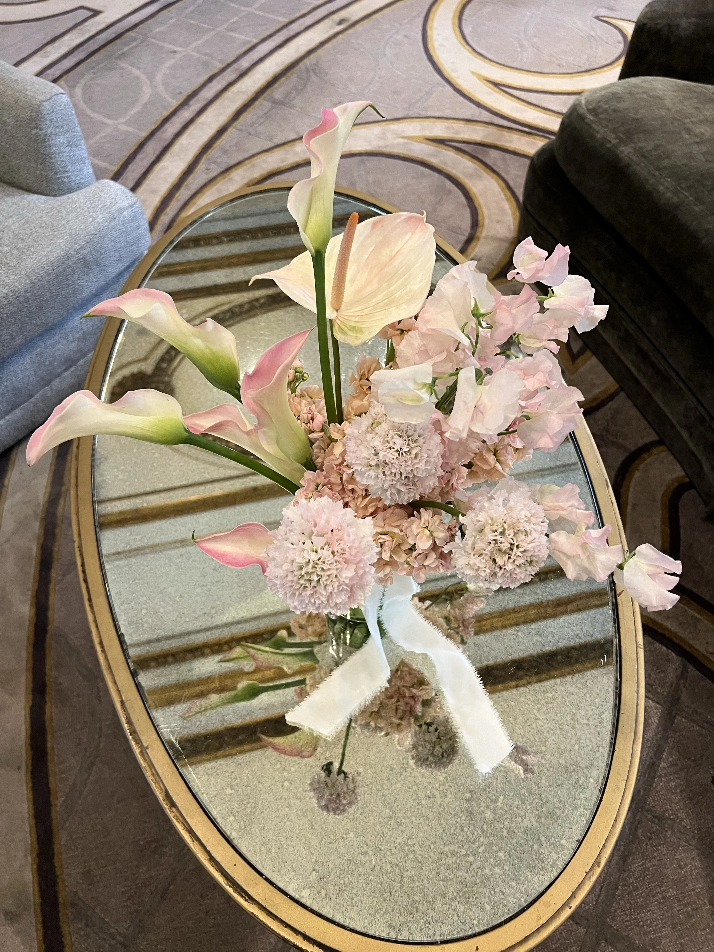 A bouquet of pink and white flowers, including calla lilies, anthuriums, and carnations, arranged on a glass-topped table with a gold-colored rim, inside a room with patterned carpet and two upholstered chairs.