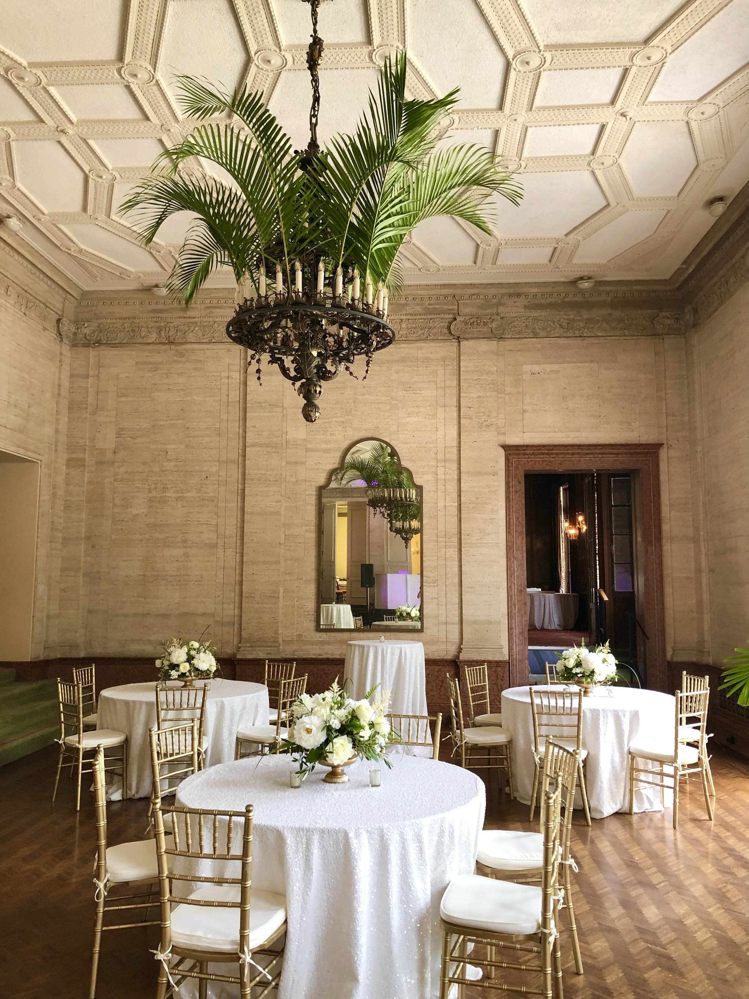 Elegant banquet room with round tables covered in white tablecloths, decorated with floral centerpieces, surrounded by gold chairs with white cushions, a large mirror on the wall, and ornate ceiling moldings.