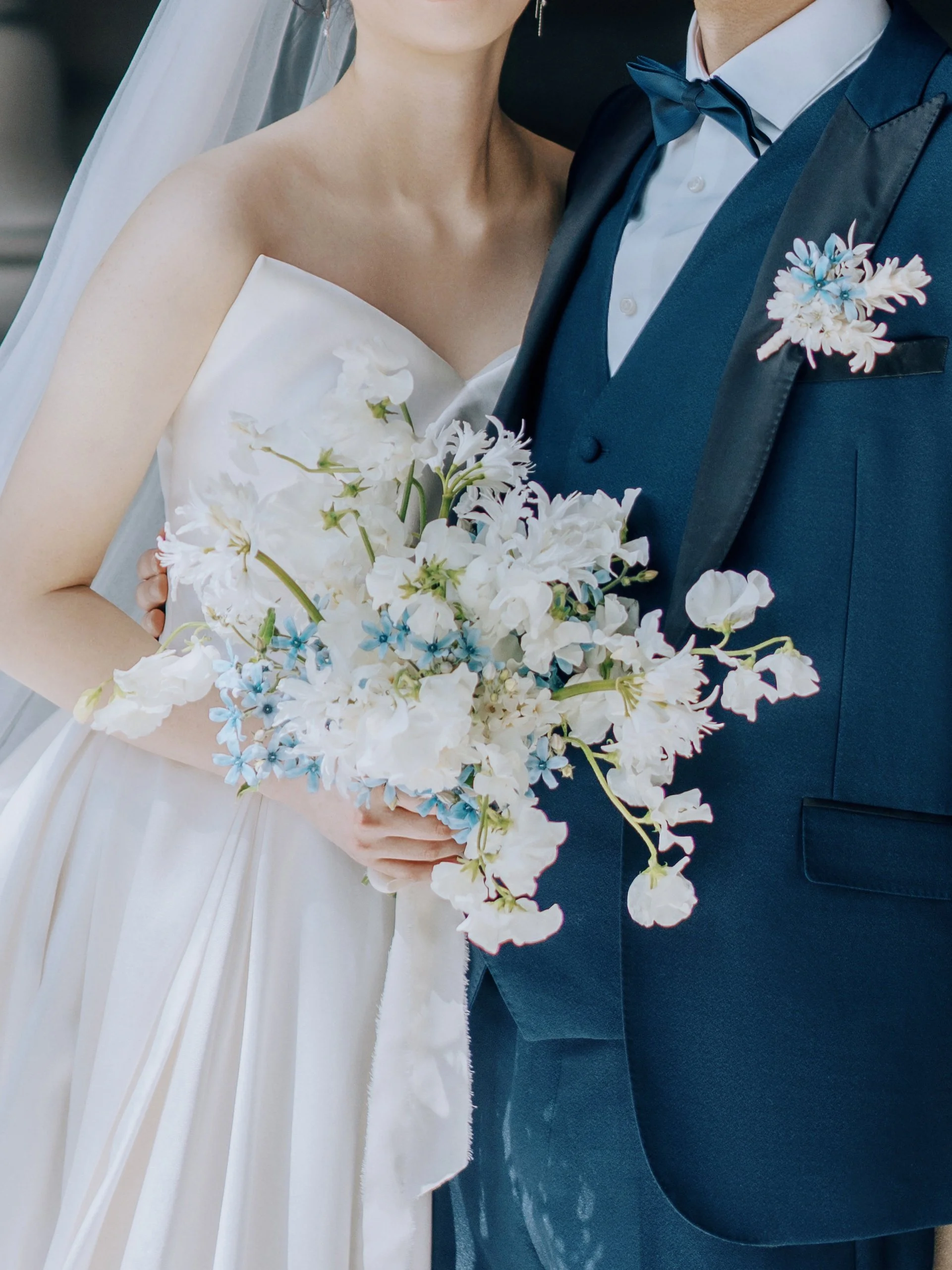 Close-up of a bride in a strapless white wedding dress holding a bouquet of white and light blue flowers standing next to a groom in a dark blue tuxedo with a bow tie, on a dark background.