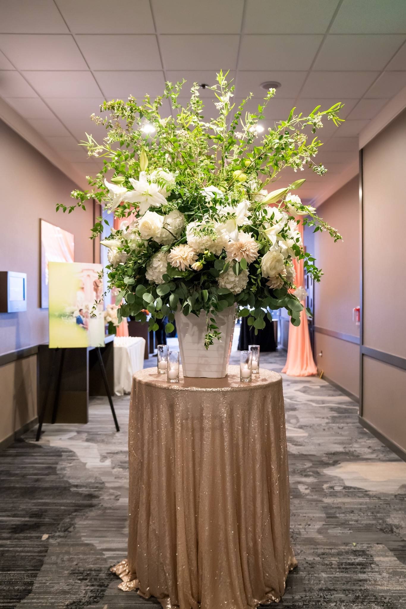 Tall floral arrangement with white lilies, roses, and greenery on a round table draped with a shimmering, gold-sequined cloth inside a decorated event hall.