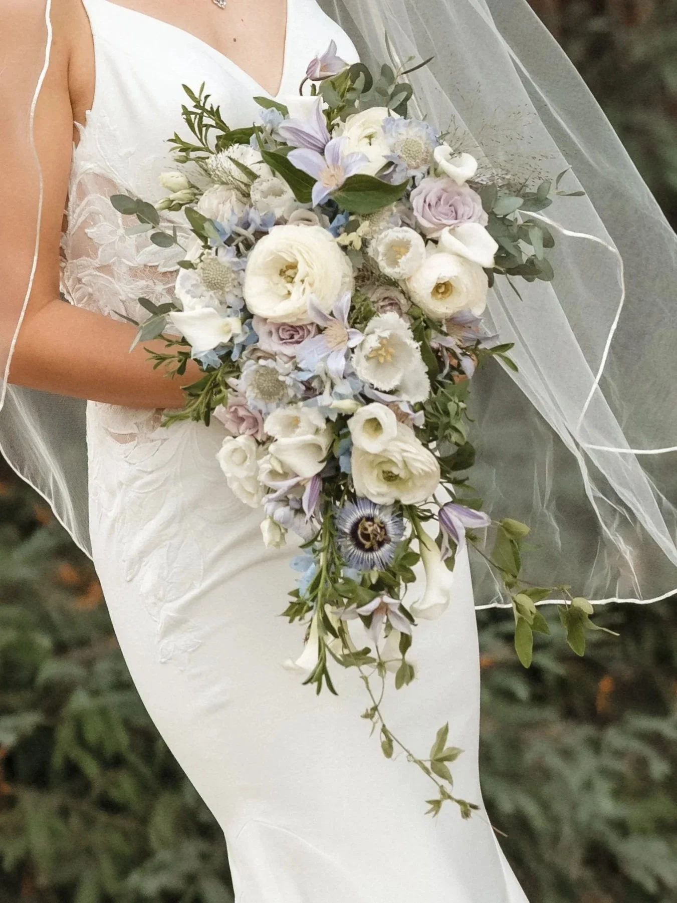 A person in a white wedding dress holding a cascading bouquet of white, pale pink, and light purple flowers with green foliage.