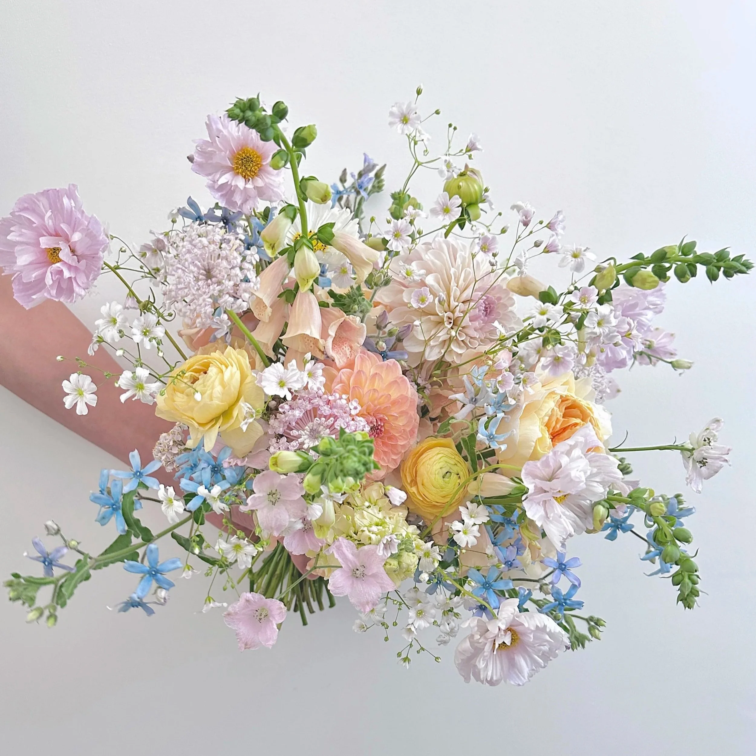 Assorted pastel-colored flowers including pink, yellow, white, and blue blooms, arranged in a bouquet with a white background.