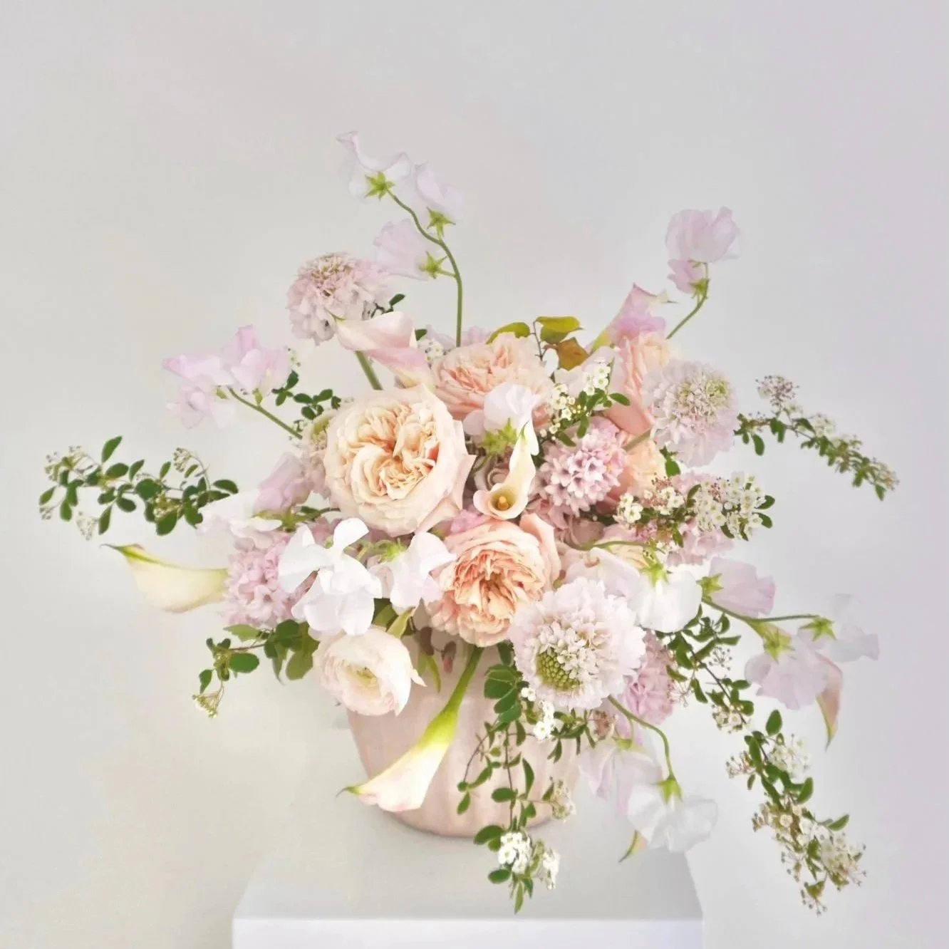 A floral arrangement in a pink vase with pink, white, and cream-colored flowers and green foliage, set against a light background.