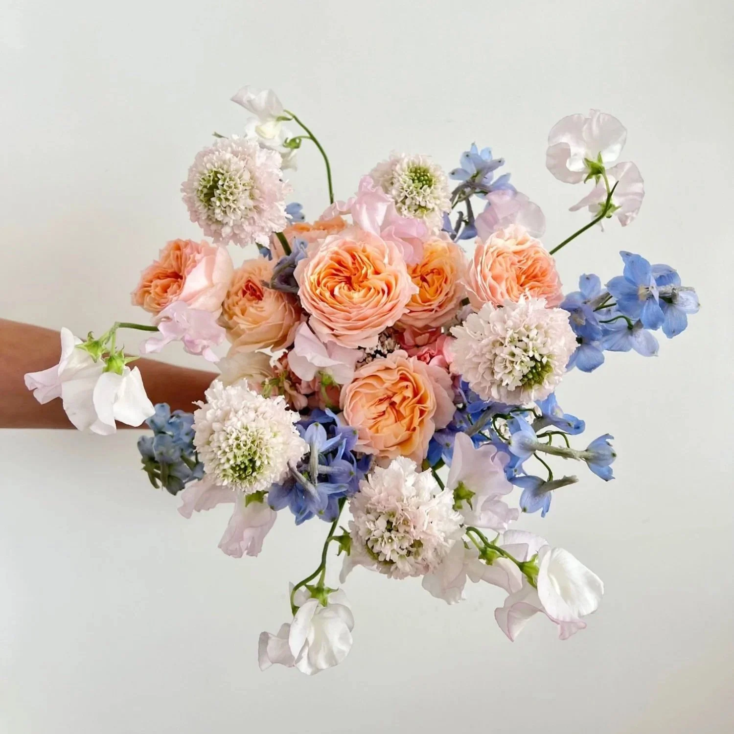 A hand holding a bouquet of assorted pastel-colored flowers, including peach roses, white sweet peas, blue delphiniums, and white scabiosa, against a plain white background.