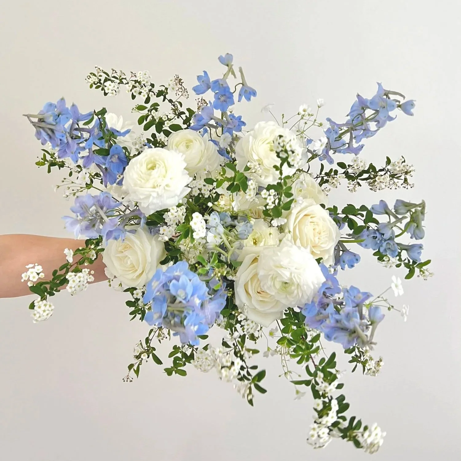 A bouquet of white roses, blue delphiniums, and small white flowers with green leaves.