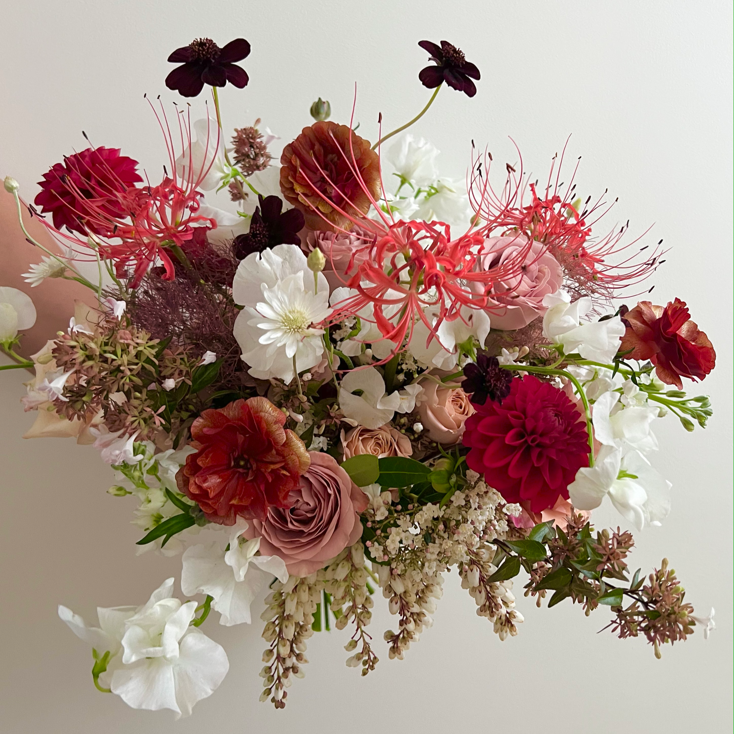 A diverse bouquet of flowers, including white, pink, deep purple, and red blossoms, arranged together against a white wall background.