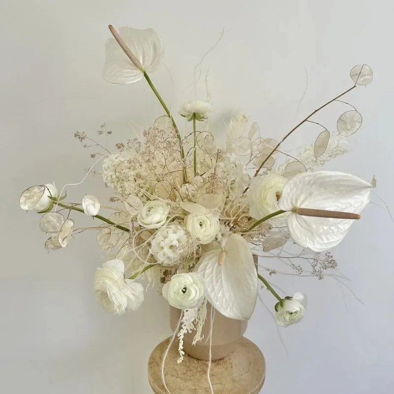 A monochromatic arrangement of white flowers and grasses in a beige vase against a plain background.