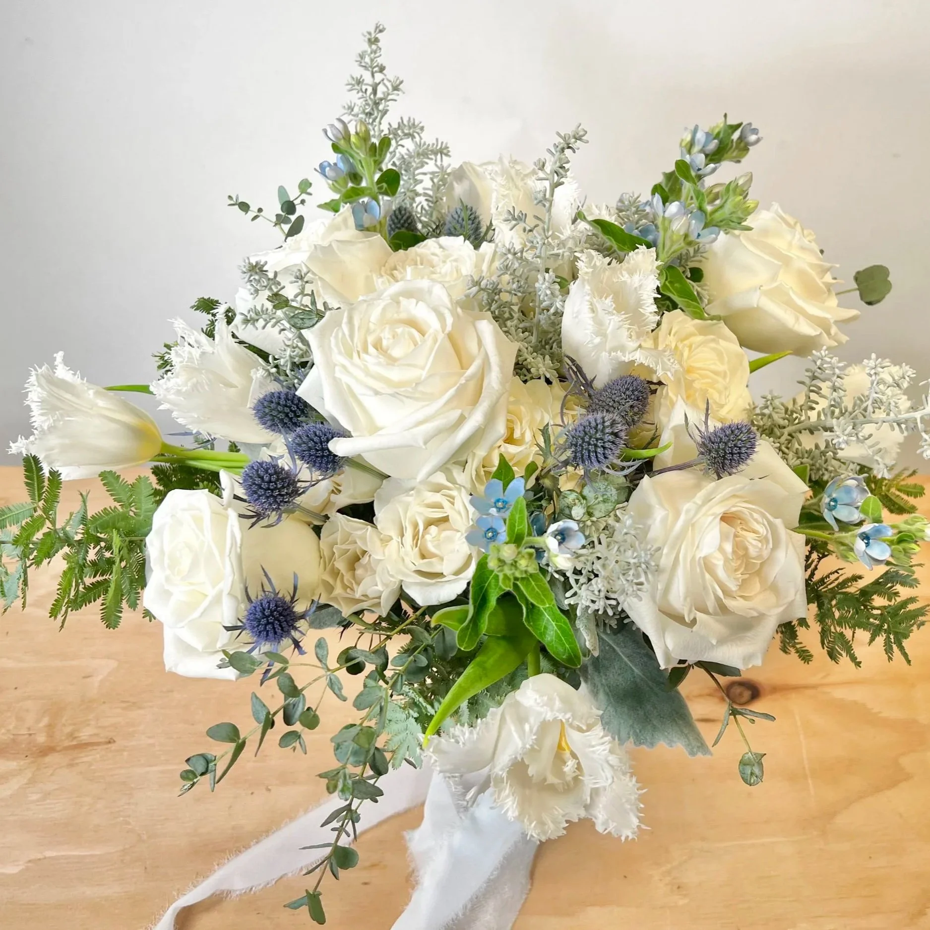 A bouquet of white roses, blue flowers, and various green and silvery foliage arranged on a wooden table.