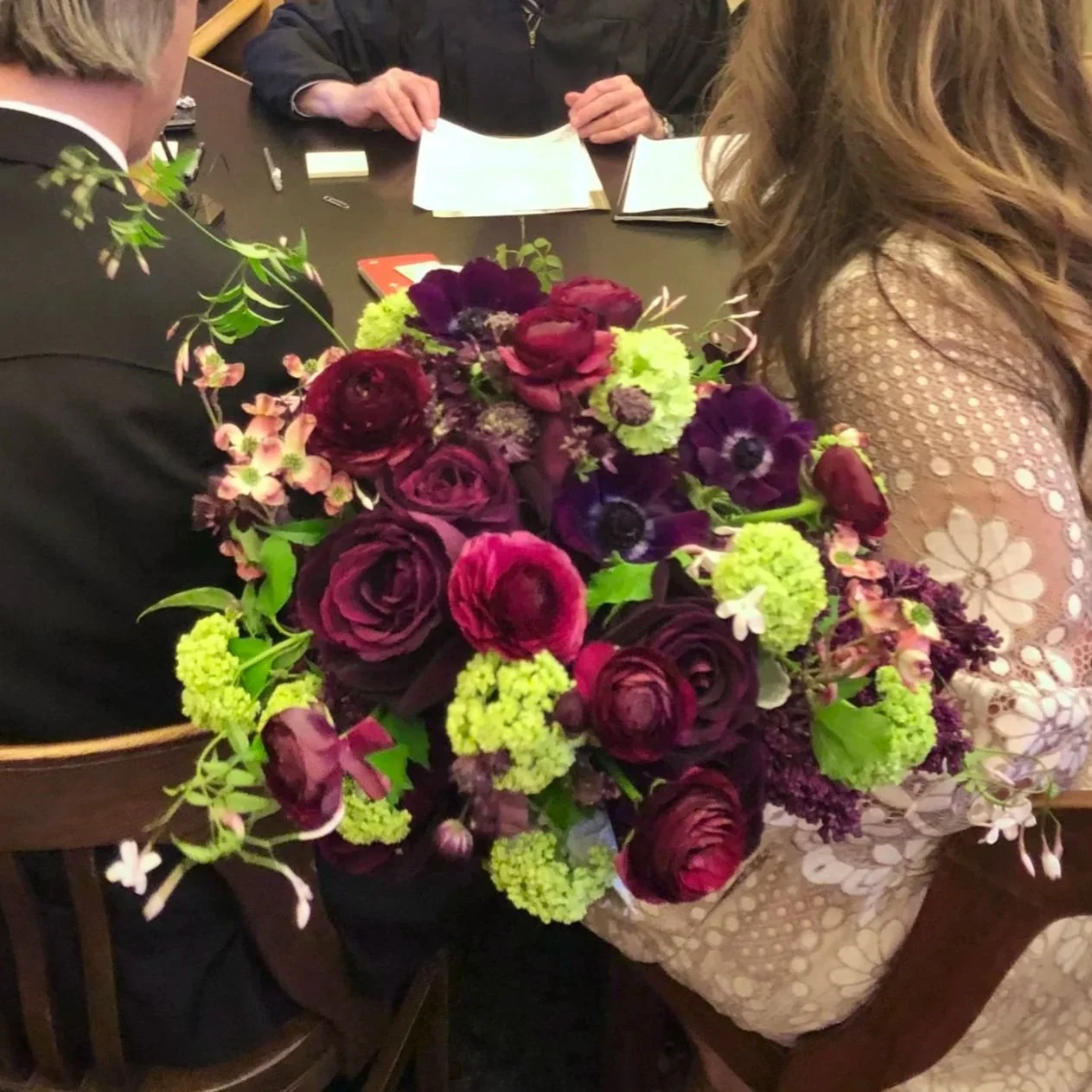 A woman sitting at a table holding a large bouquet of dark purple and green flowers, with people in the background reading notebooks.