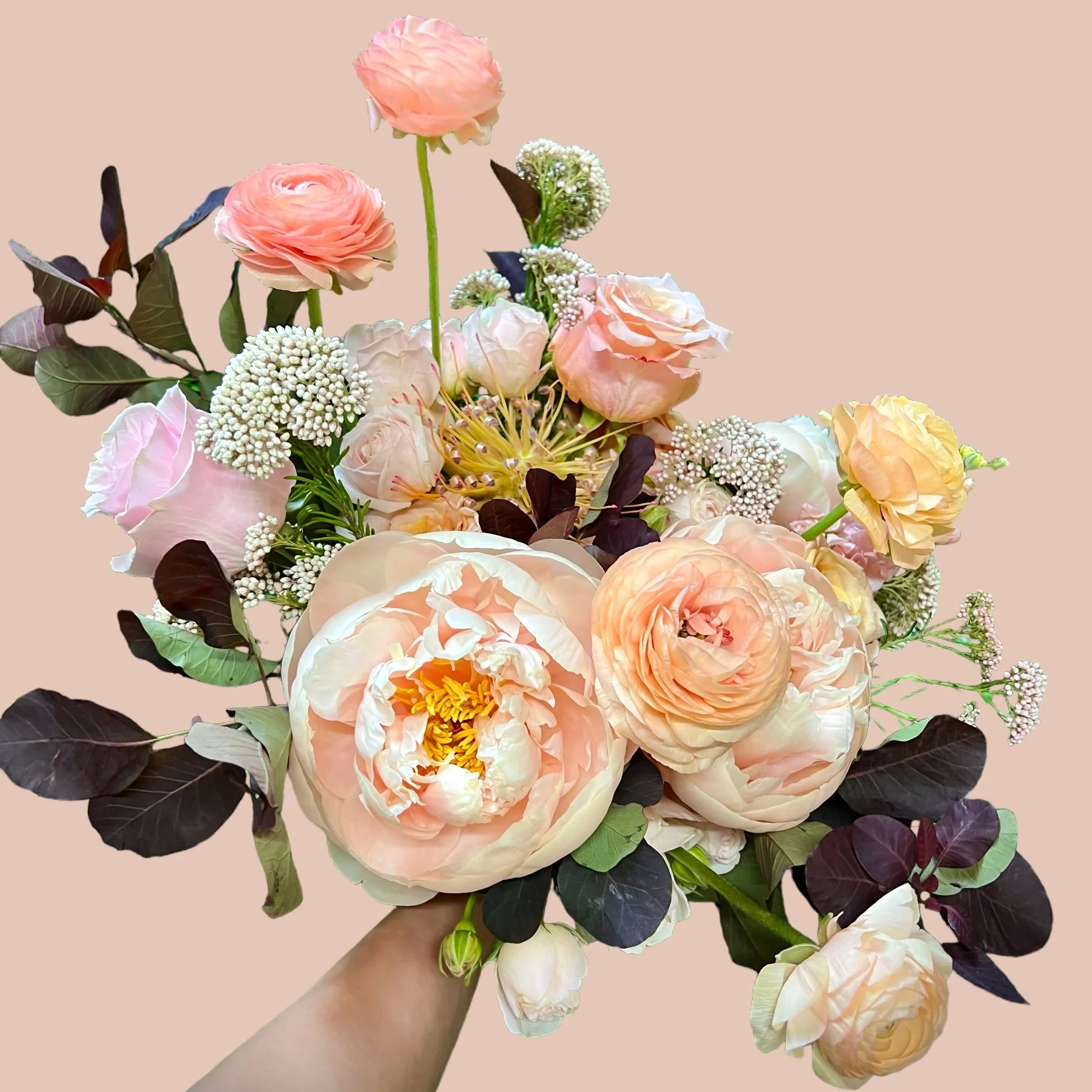 Hand holding a bouquet of soft pink, cream, and white peonies, ranunculus, and small white filler flowers against a pale beige background.