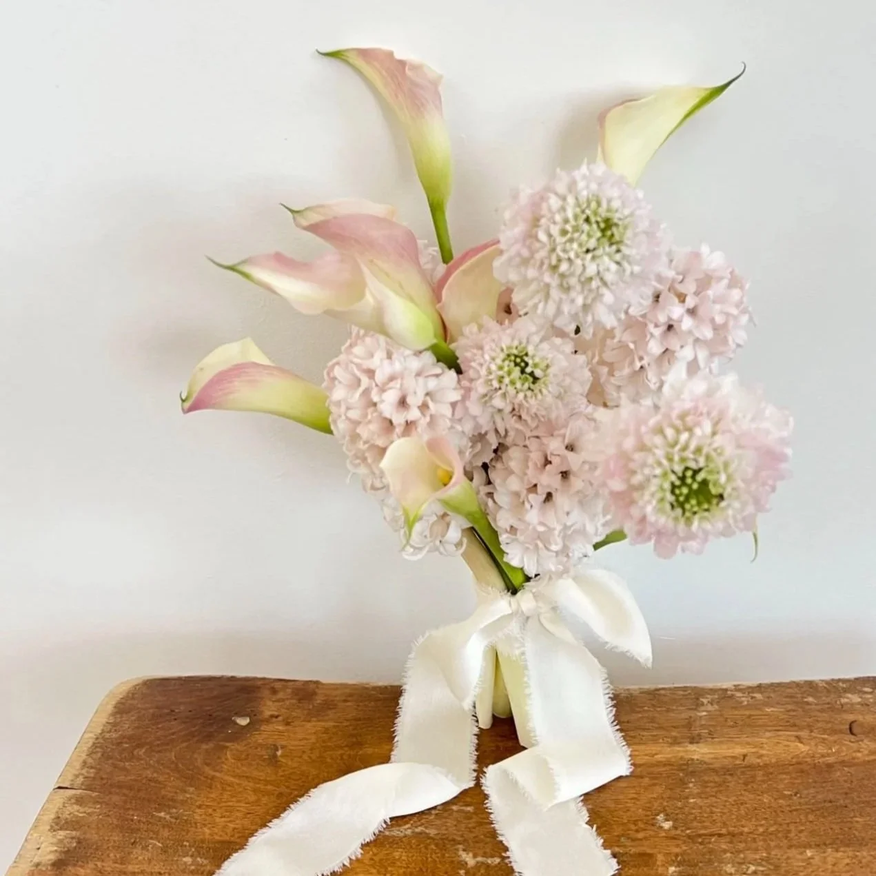 A bouquet of pale pink and white flowers with ribbon, placed on a wooden surface against a white background.