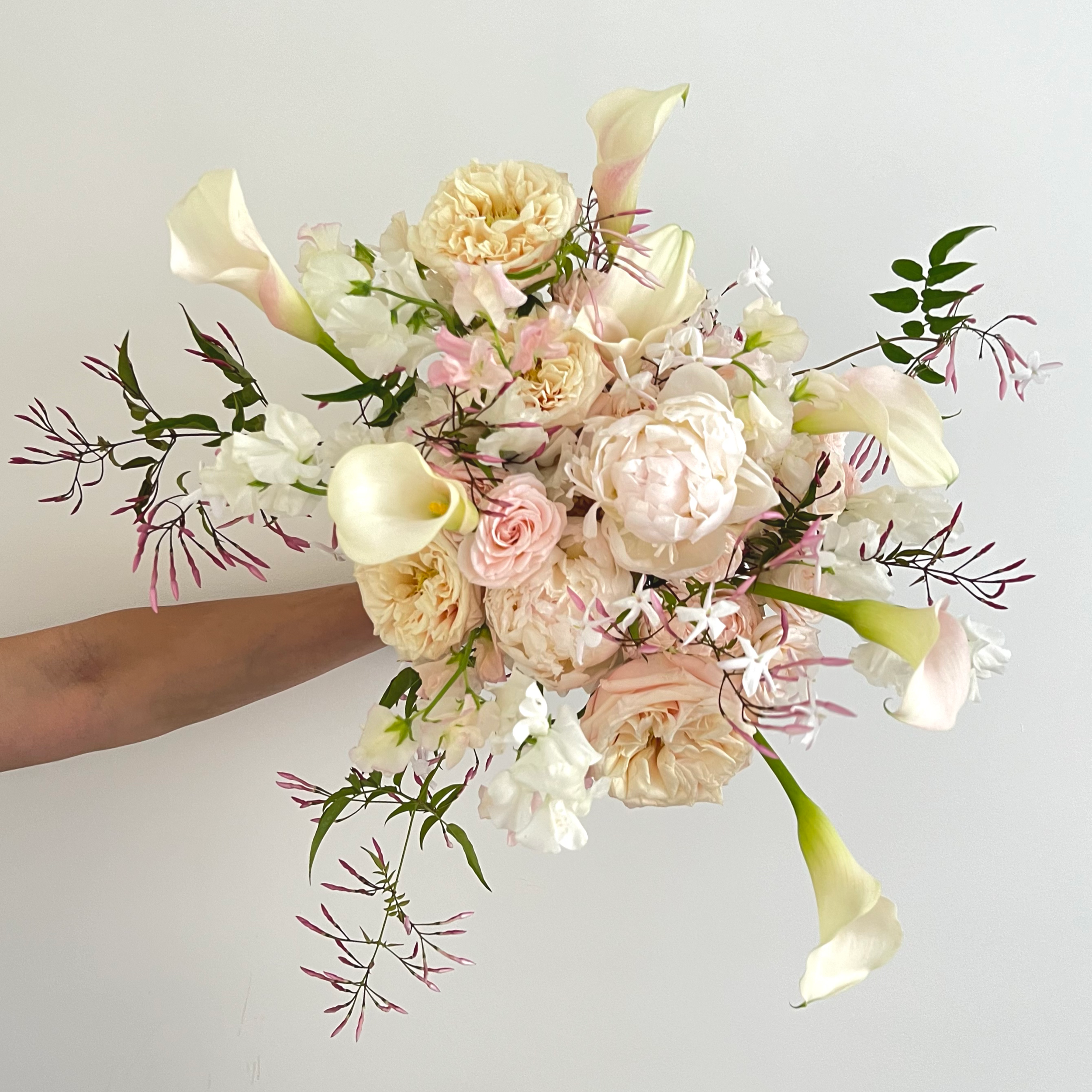 Arrangement of cream, white, and pink flowers, including calla lilies and roses, with green and purple foliage, against a plain white background.