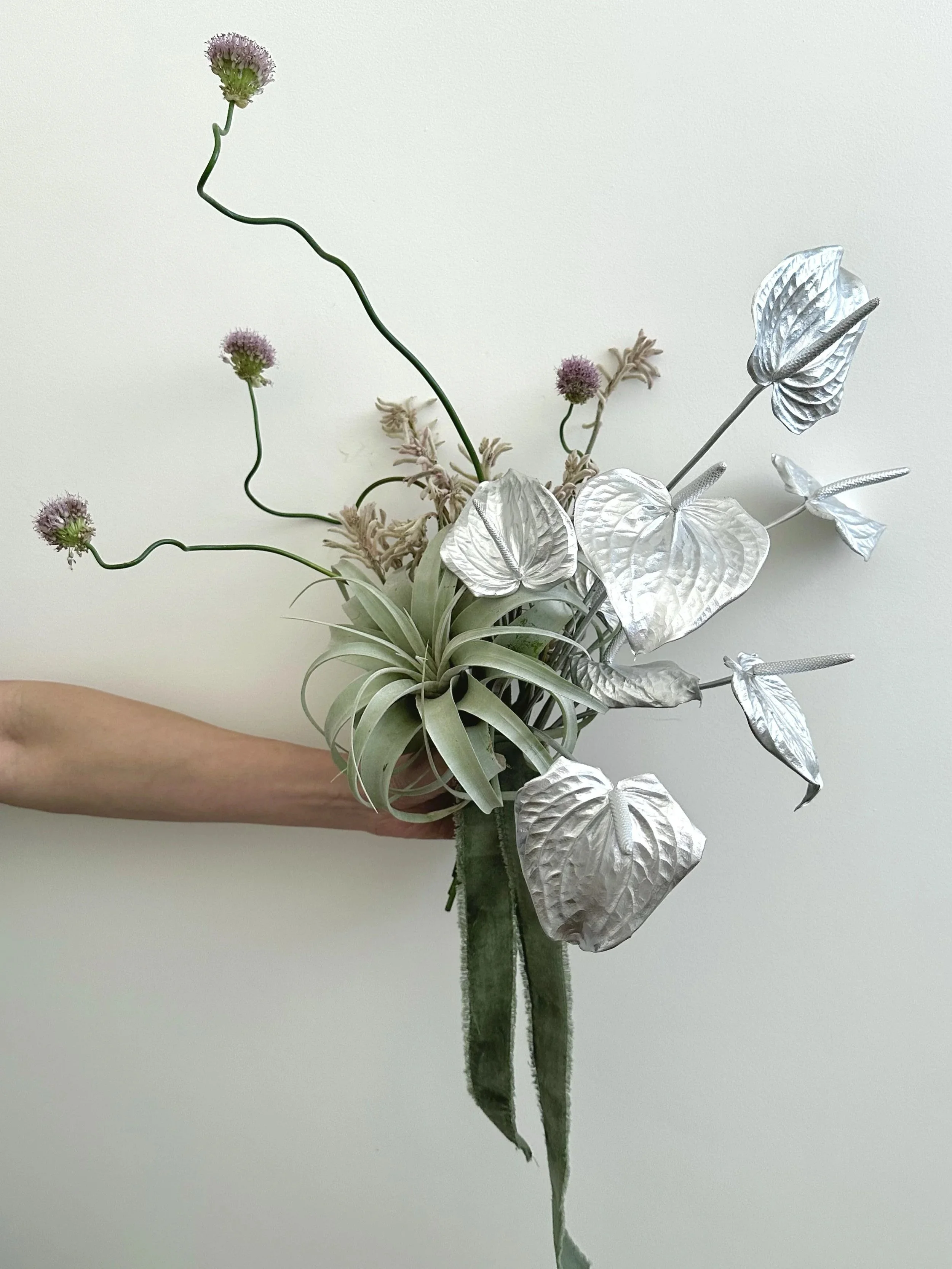 A hand holding a floral arrangement with greenish-gray leaves, purple flowers, and metallic silver leaves and stems against a plain white background.