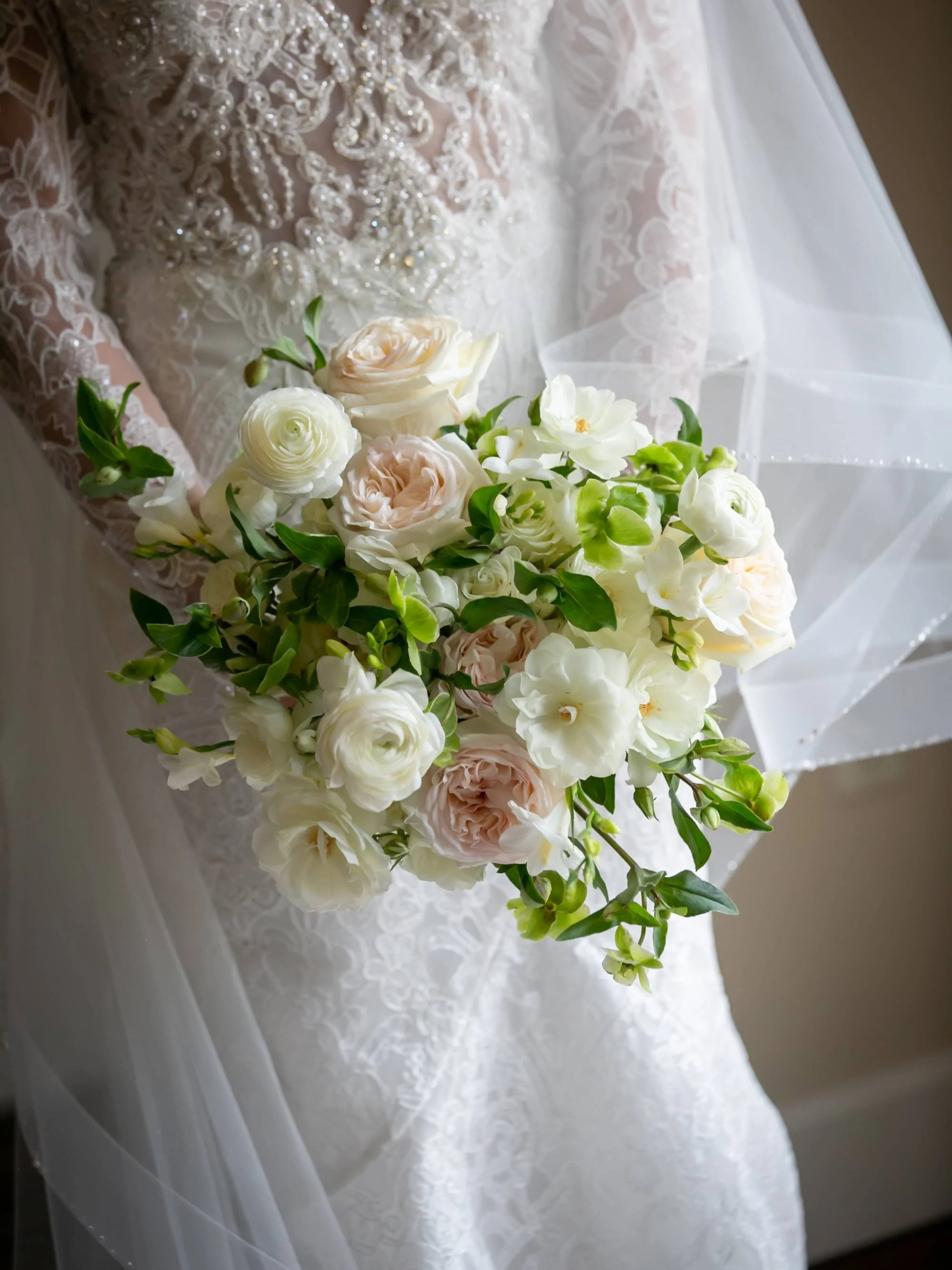 Close-up of a bouquet of white and cream roses and green leaves held against a lace wedding dress with intricate beadwork and embroidery.