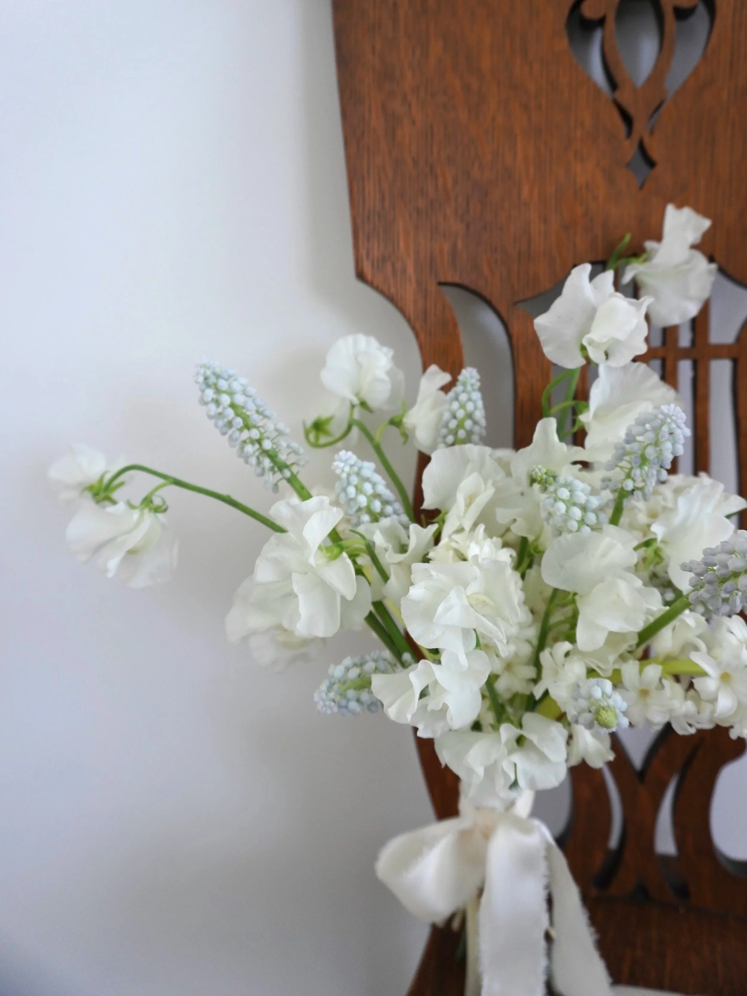 A bouquet of white flowers, including sweet peas and possibly grape hyacinths, sits on a wooden chair against a plain white wall.