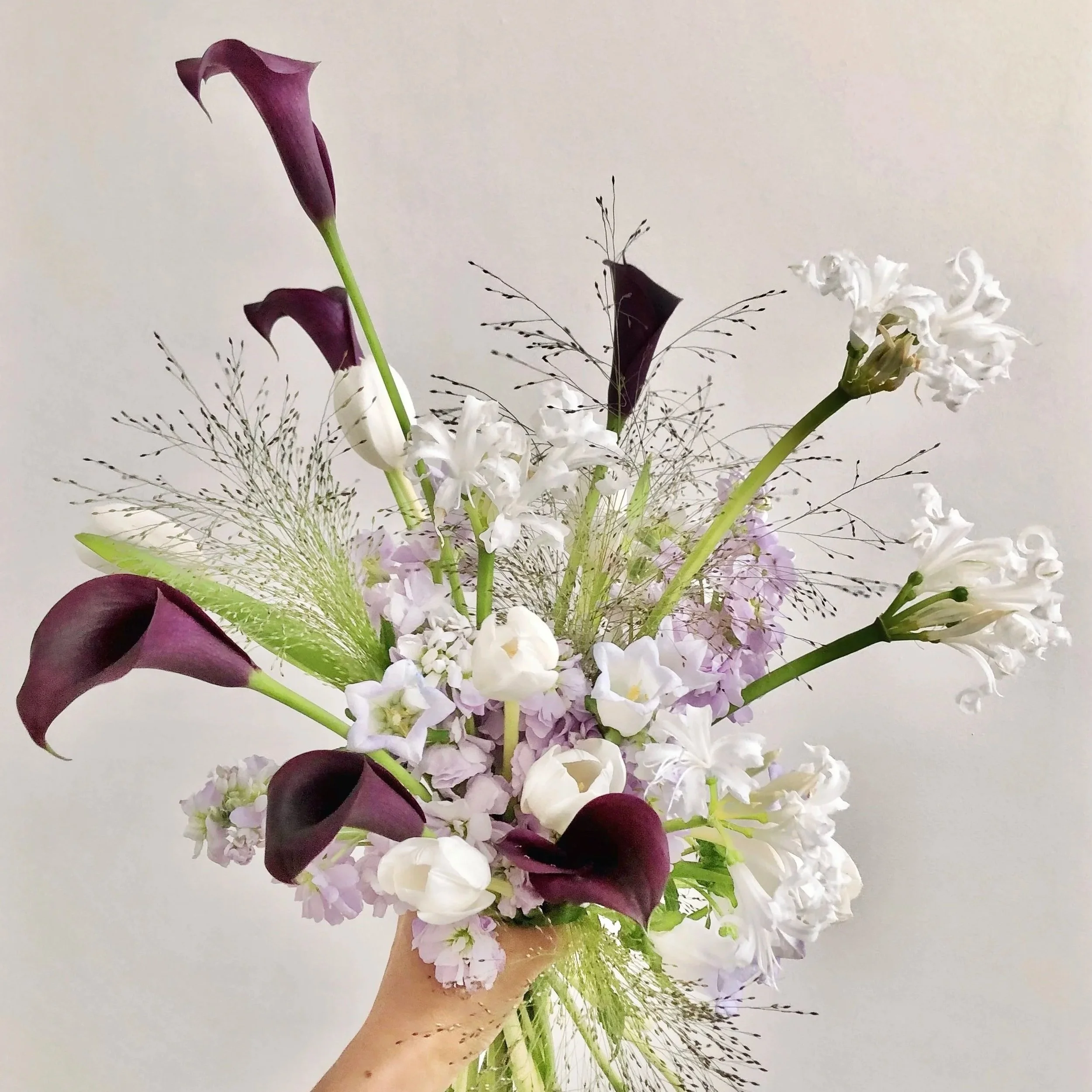A person holding a bouquet of mixed flowers including purple calla lilies, white orchids, and other white and purple blooms, against a plain wall background.