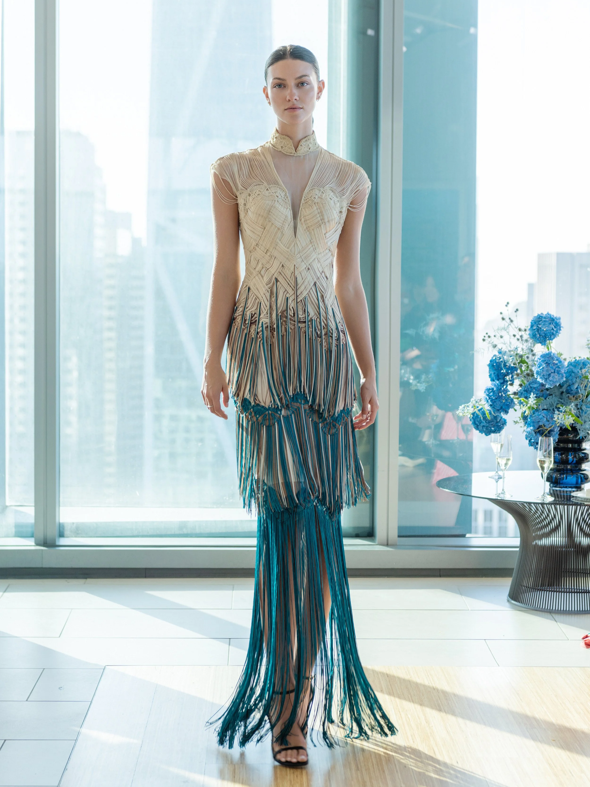 A woman wearing a long, colorful, fringed dress with a high neckline, standing indoors near large glass windows with a city skyline in the background, next to a round table with a vase of blue flowers and two champagne glasses.