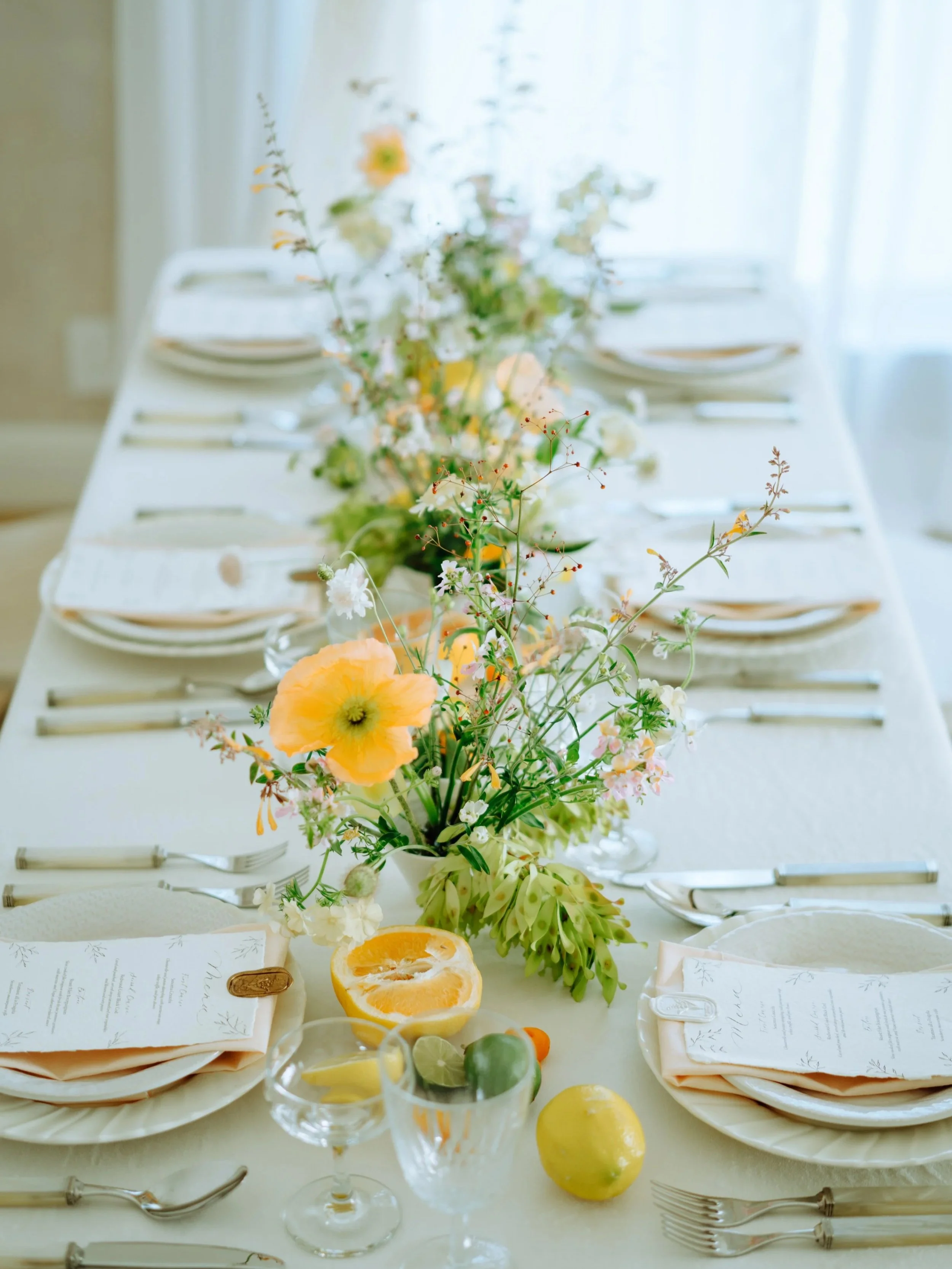 Elegant dining table with a floral centerpiece, sliced citrus fruits, glassware, silverware, and menu cards, set for a formal meal in bright natural light.