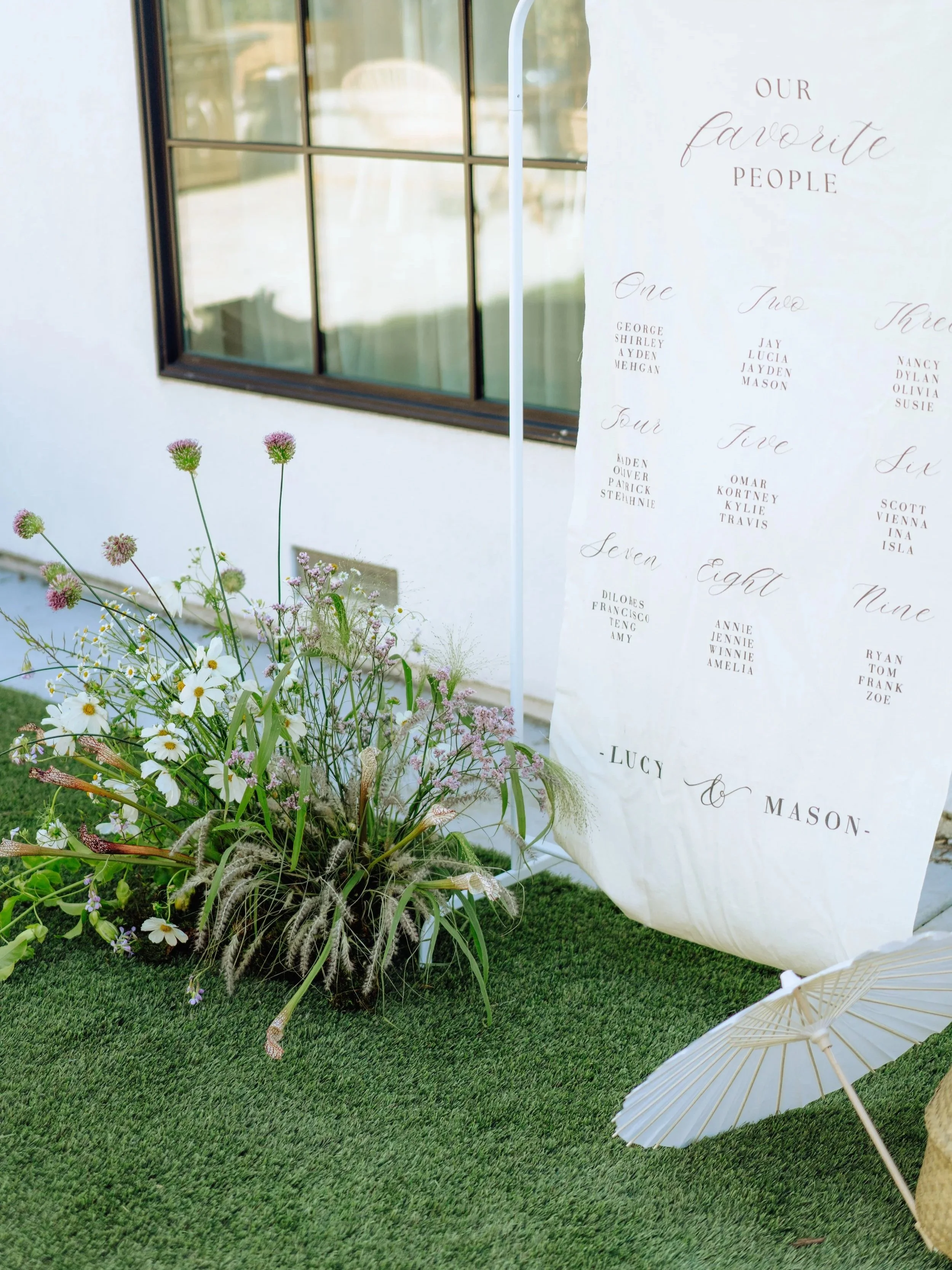 A floral display with white daisies and pink globe amaranth flowers in front of a white fabric backdrop with wedding guest names and table numbers. There is a window with black frames on the wall behind the display and a white parasol lying on the gr