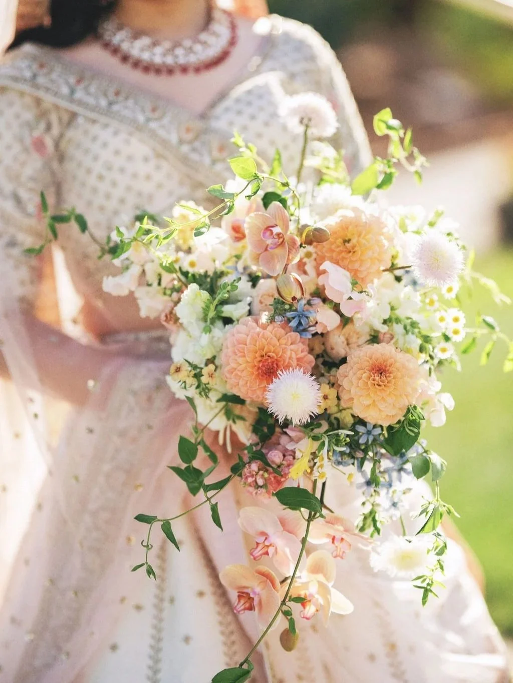 A woman in traditional Indian attire holding a bouquet of pastel-colored flowers, including dahlias, orchids, and daisies, outdoors during the daytime.