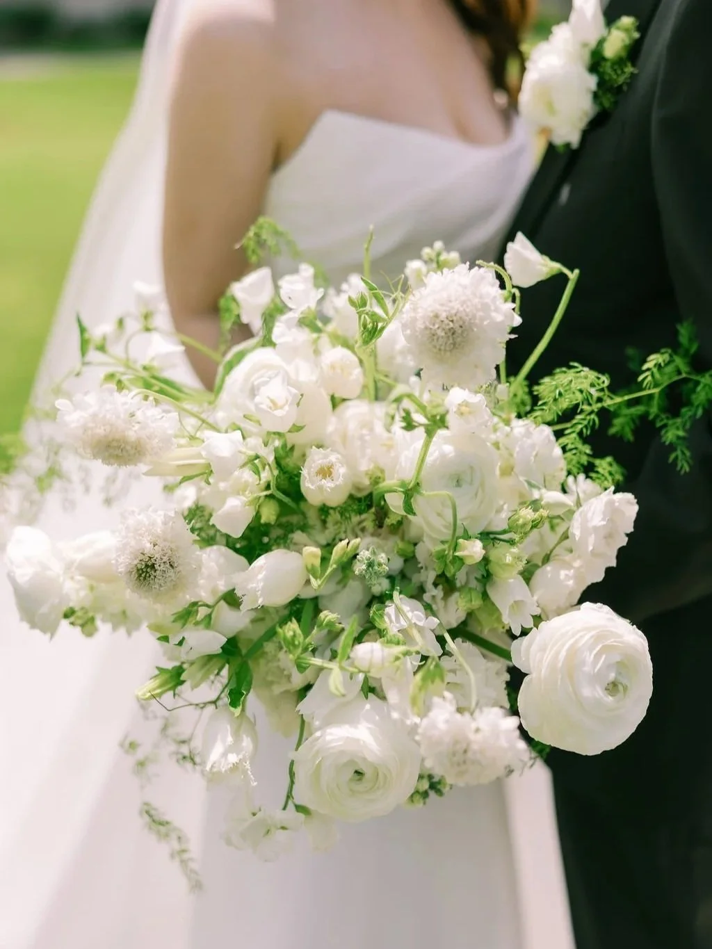 Close-up of a bride holding a white floral bouquet, with a groom in a black suit, at a wedding outdoors.