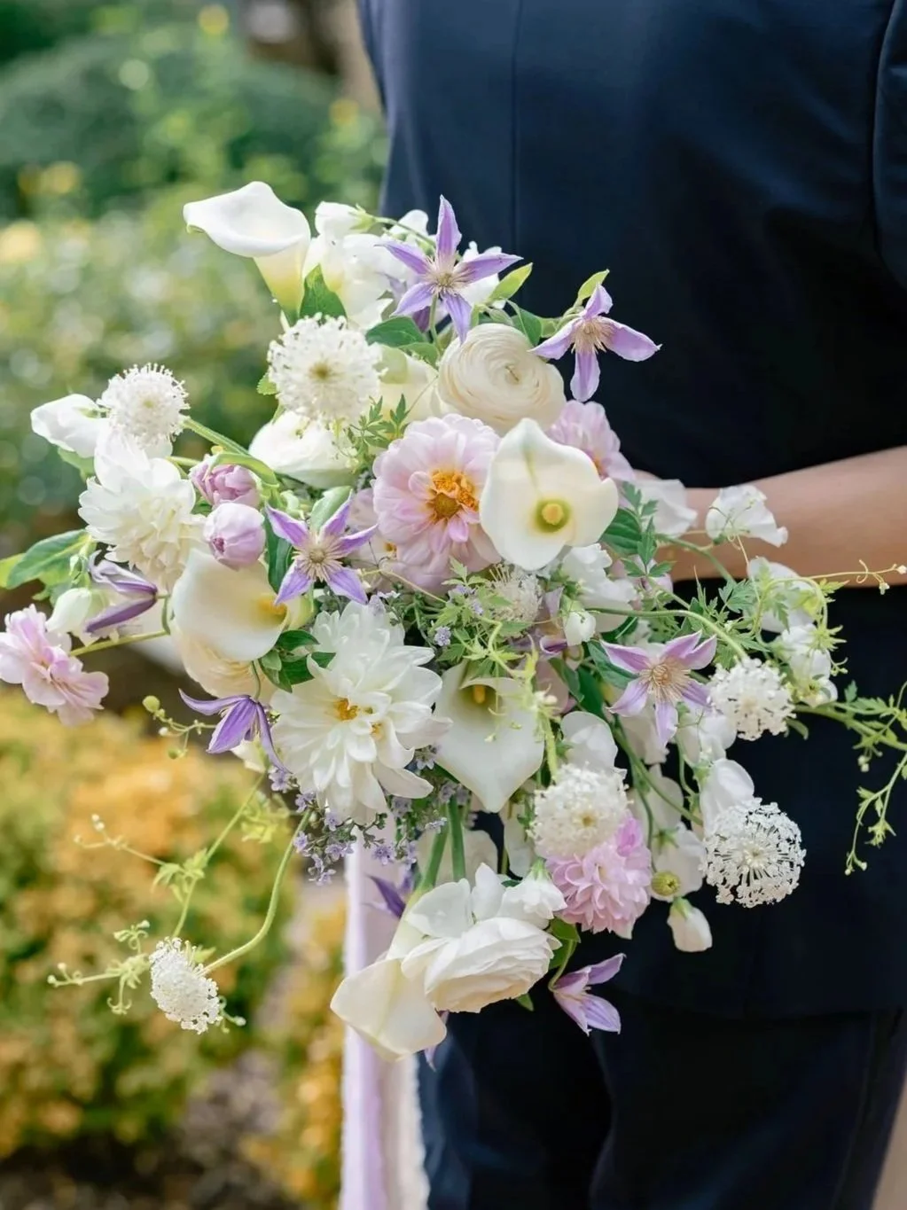 Person in dark clothing holding a bouquet of white and pastel-colored flowers outdoors.