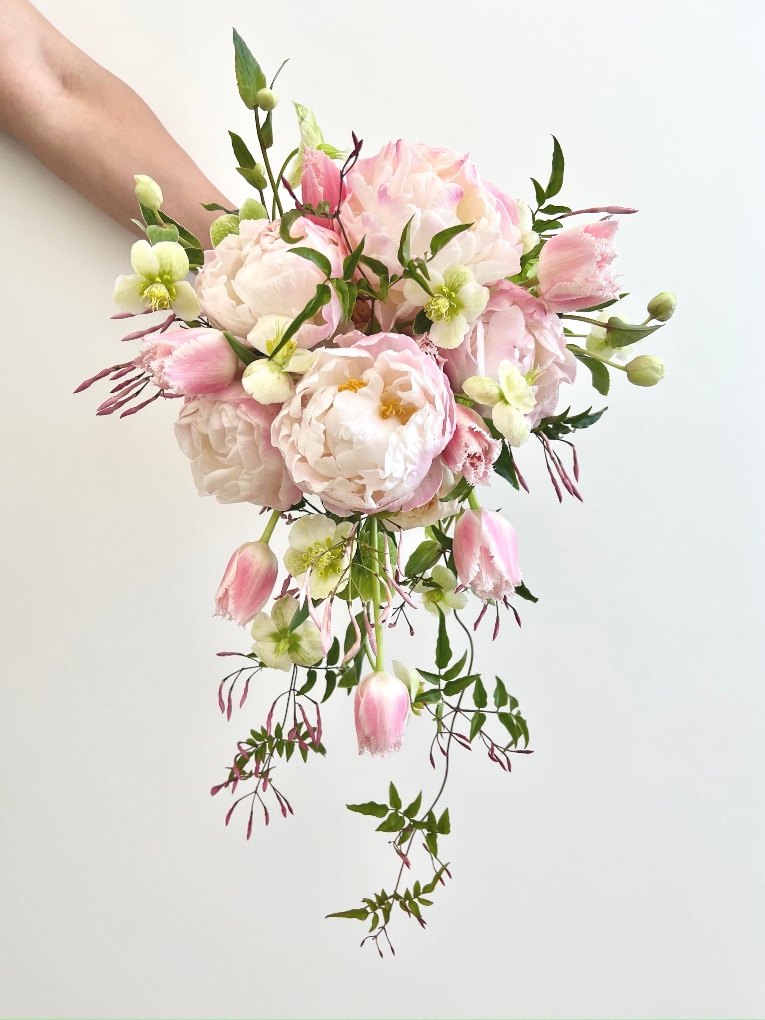 A cascading bouquet of pink and white flowers with green leaves, including peonies, tulips, and garden roses, against a plain light background.