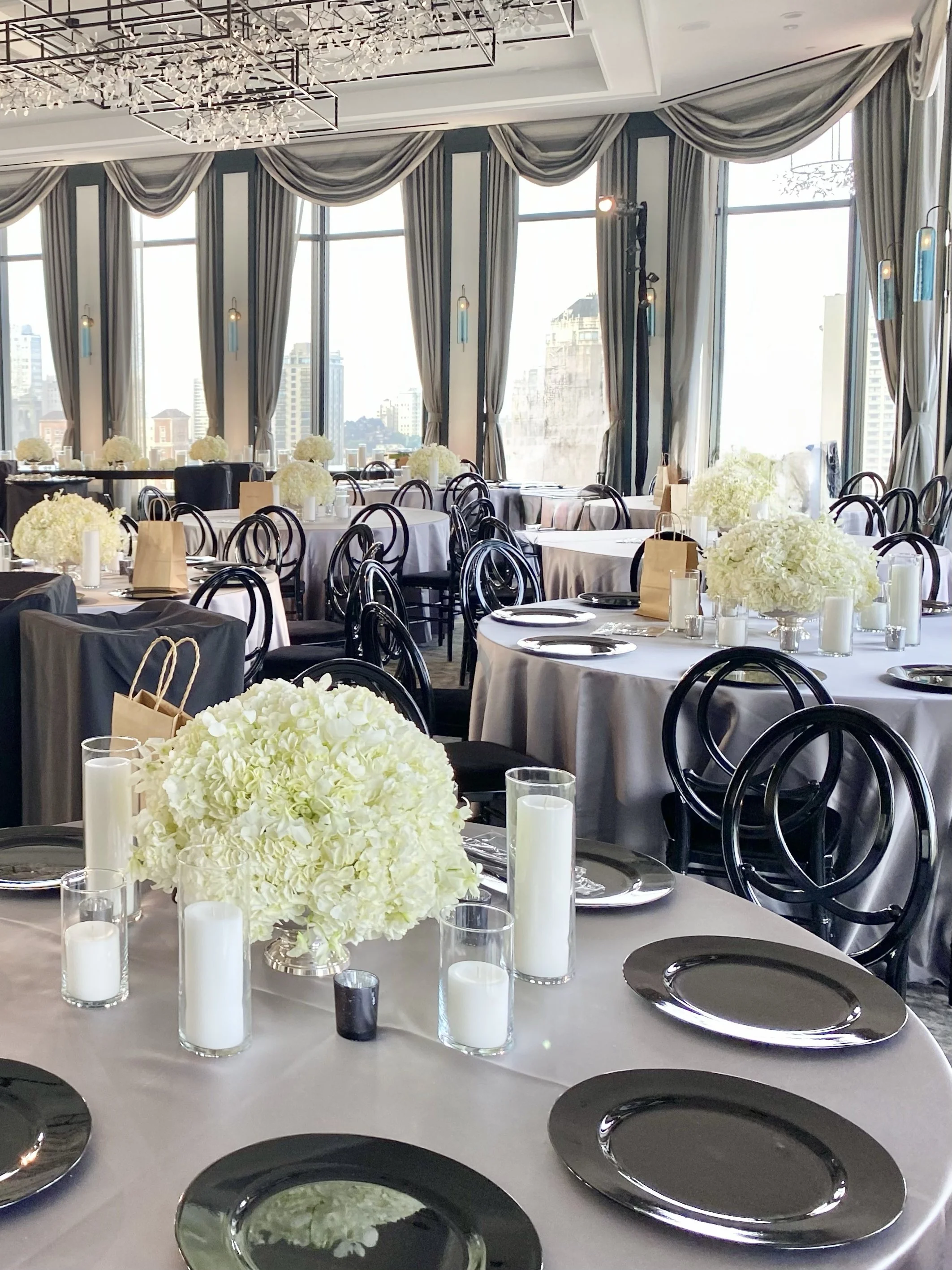 Elegant banquet hall with large windows, draped curtains, round tables with white tablecloths, decorated with white floral centerpieces, candles, black plates, and black chairs, set up for a formal event.