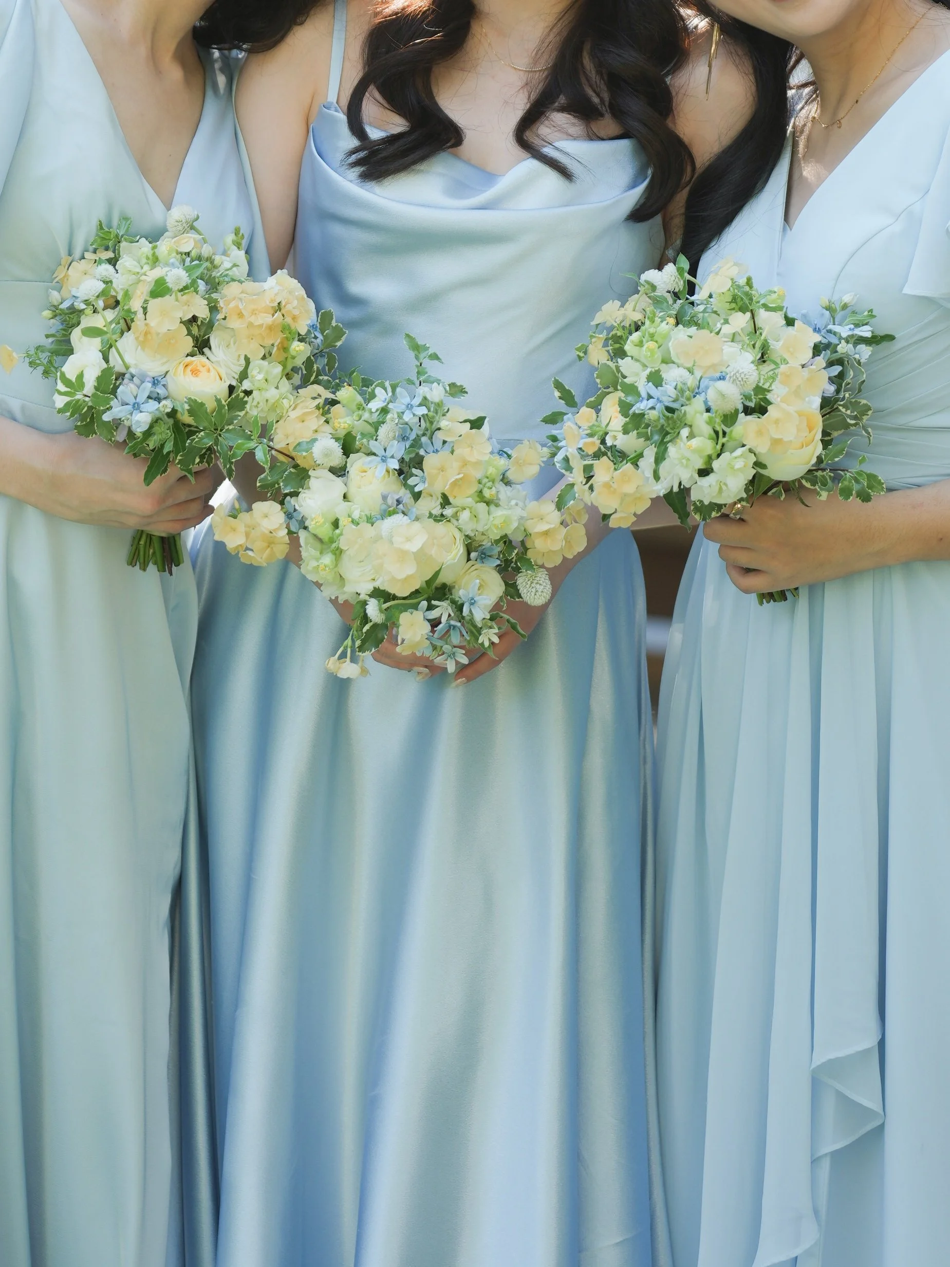 Three women in light blue dresses holding floral bouquets, standing close together.