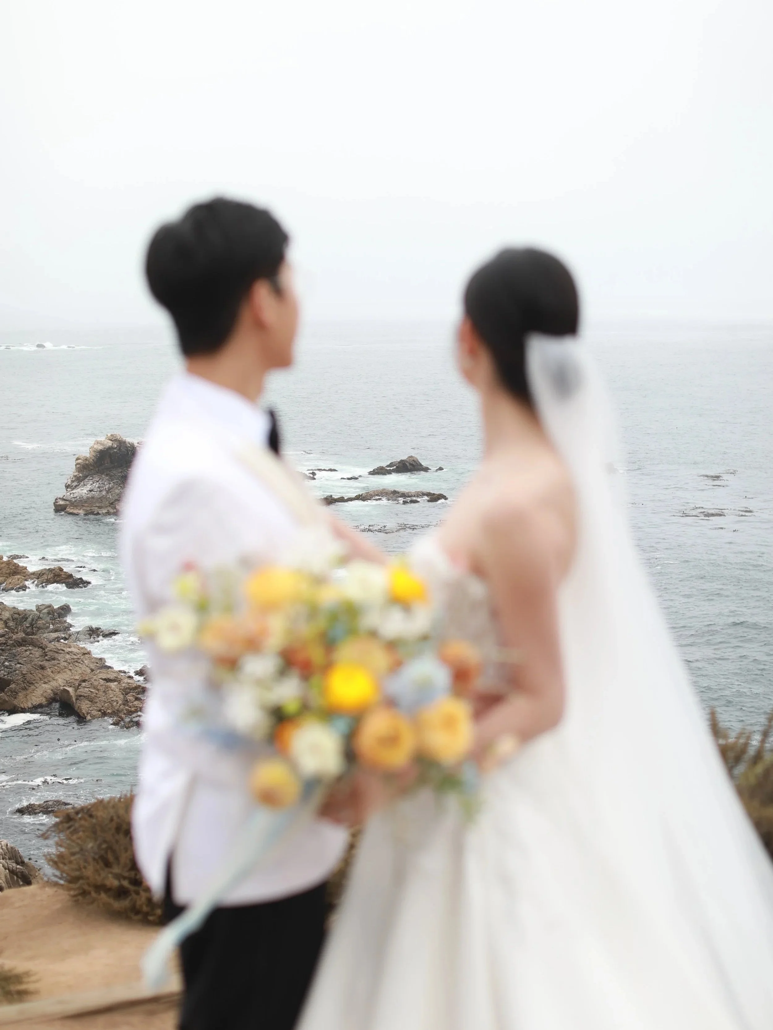 A blurred photo of a bride and groom standing on a rocky shoreline with ocean waves in the background during their wedding.