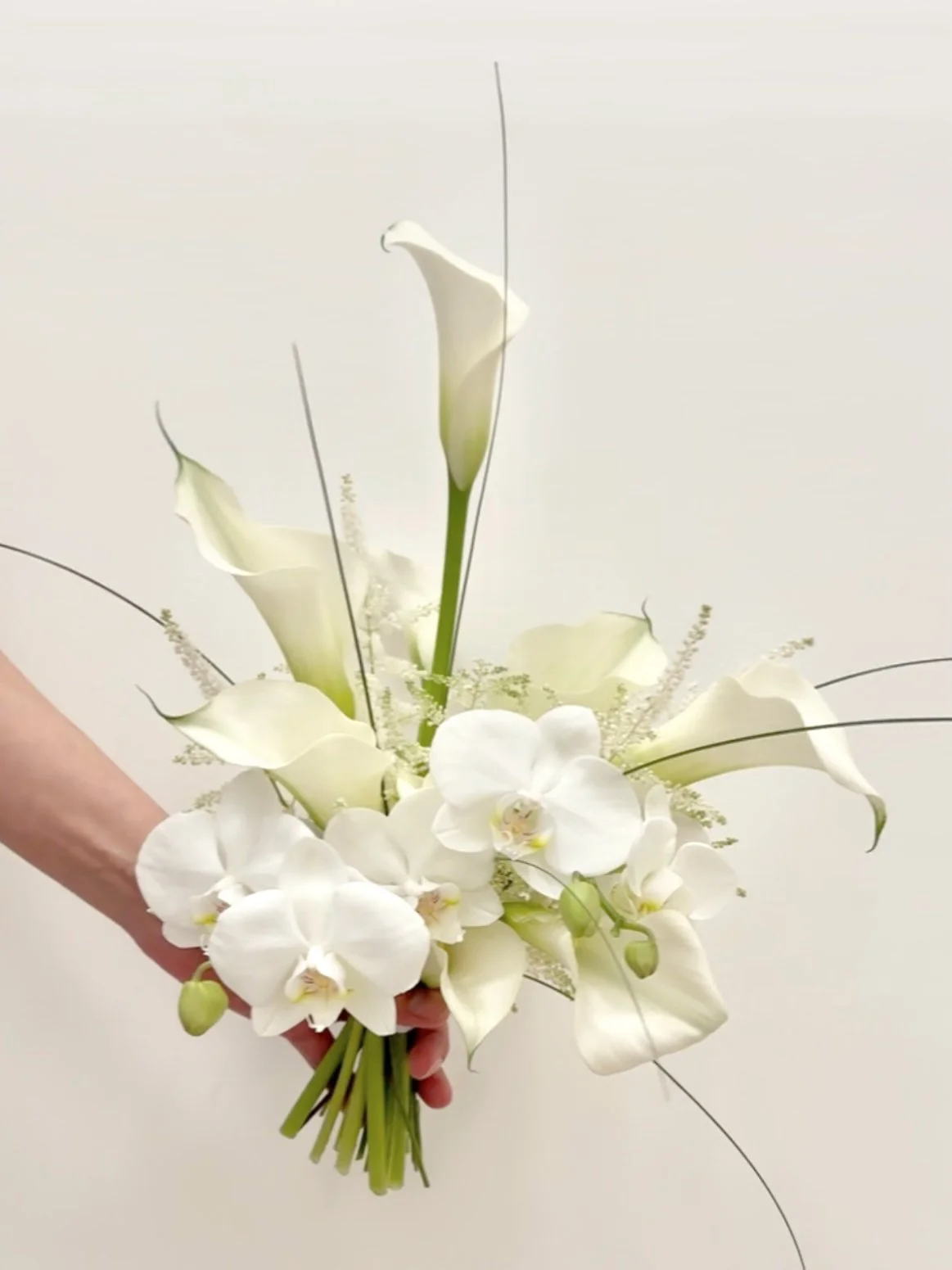 A hand holding a bouquet of white calla lilies and white orchids against a plain background.