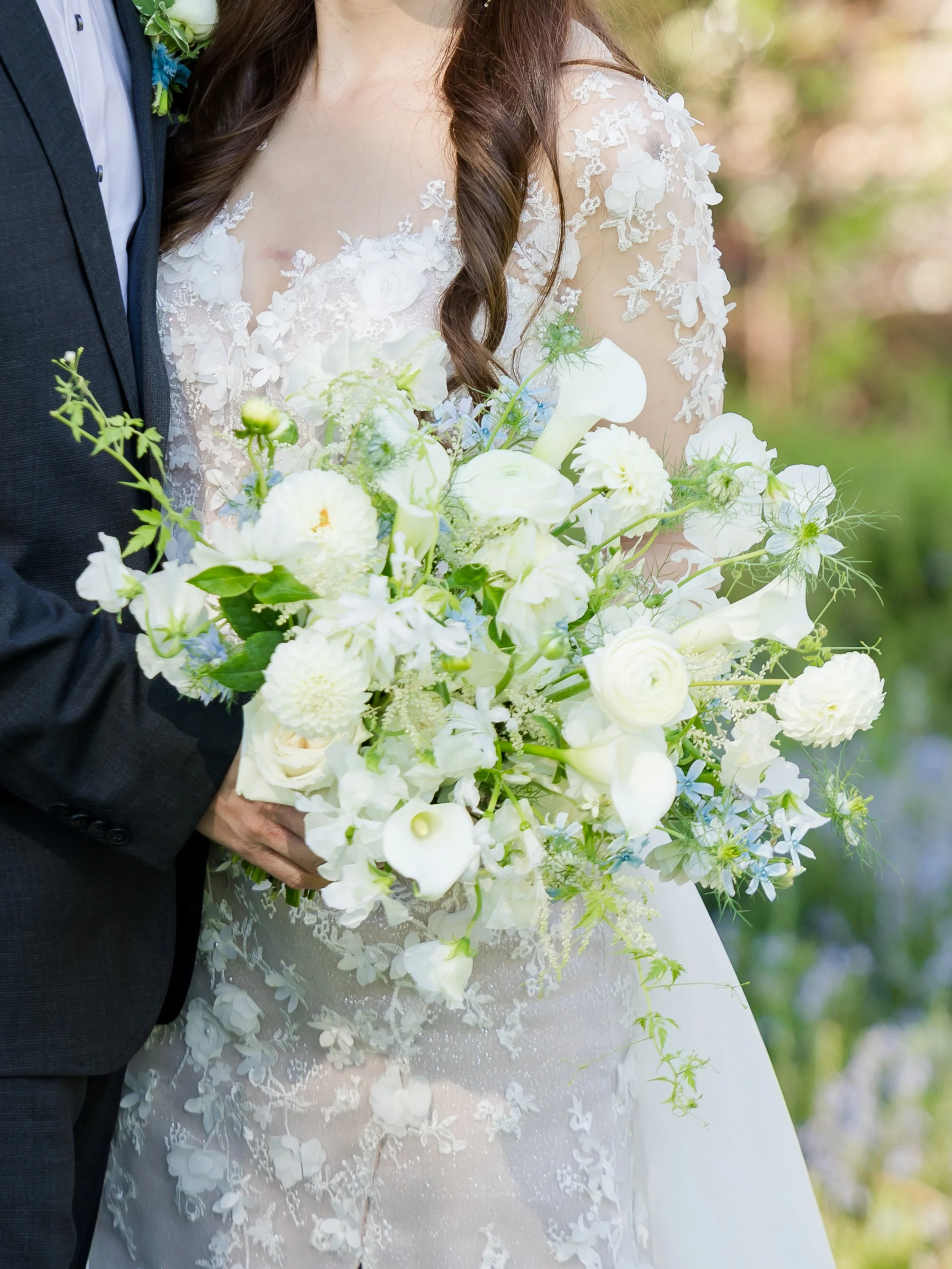 Close-up of a bride and groom holding a bouquet of white flowers on their wedding day, with the bride wearing a lace wedding dress and the groom in a dark suit.