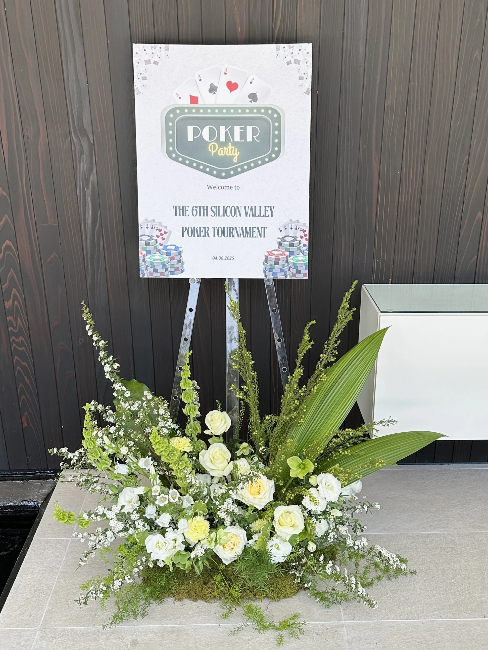 A floral arrangement in front of a sign for a poker party at the 6th Silicon Valley Poker Tournament, with a dark wooden wall background.