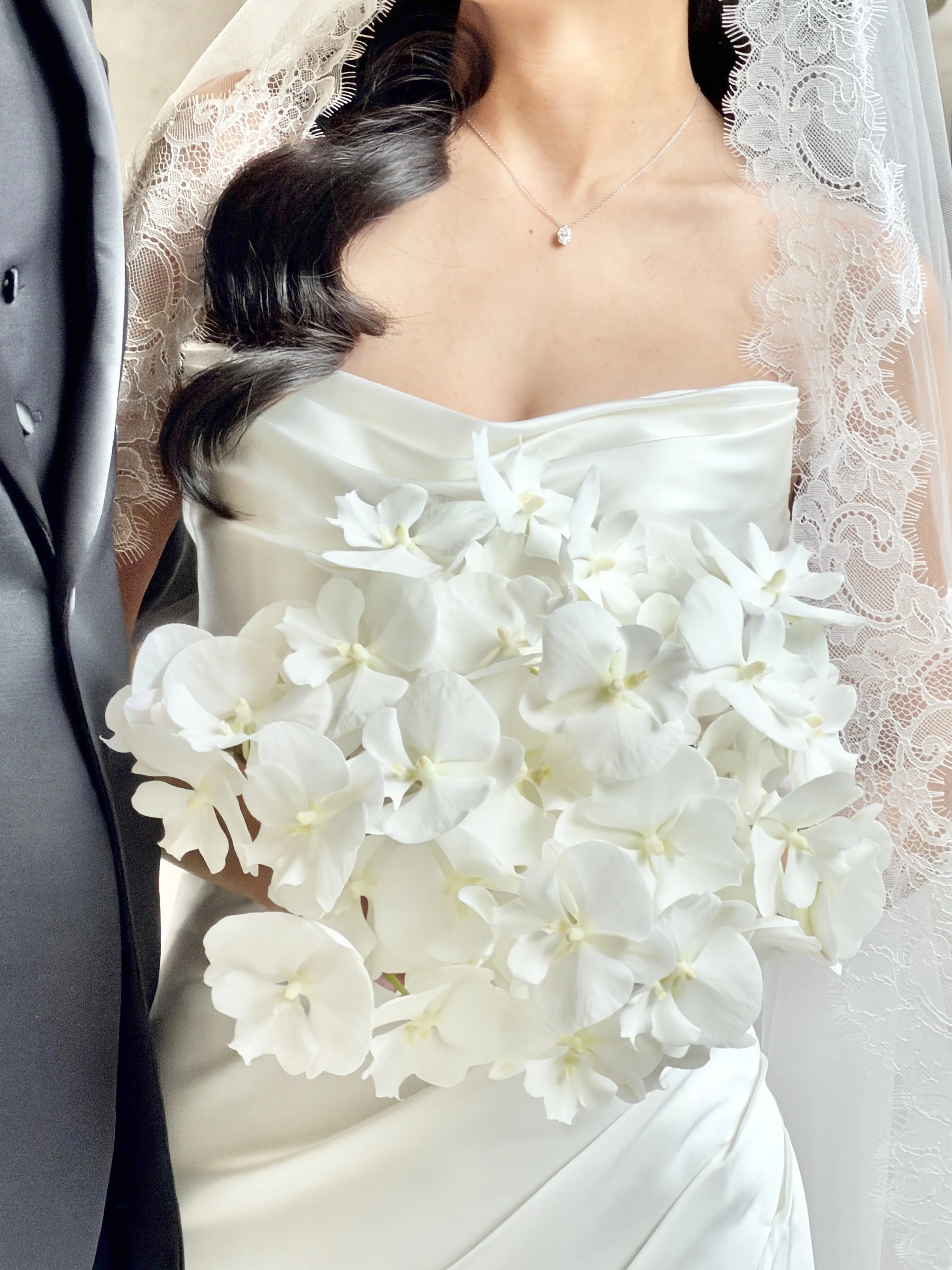 A bride in a white wedding dress with lace details, lying on a surface with white orchids placed on her dress, and wearing a delicate necklace.