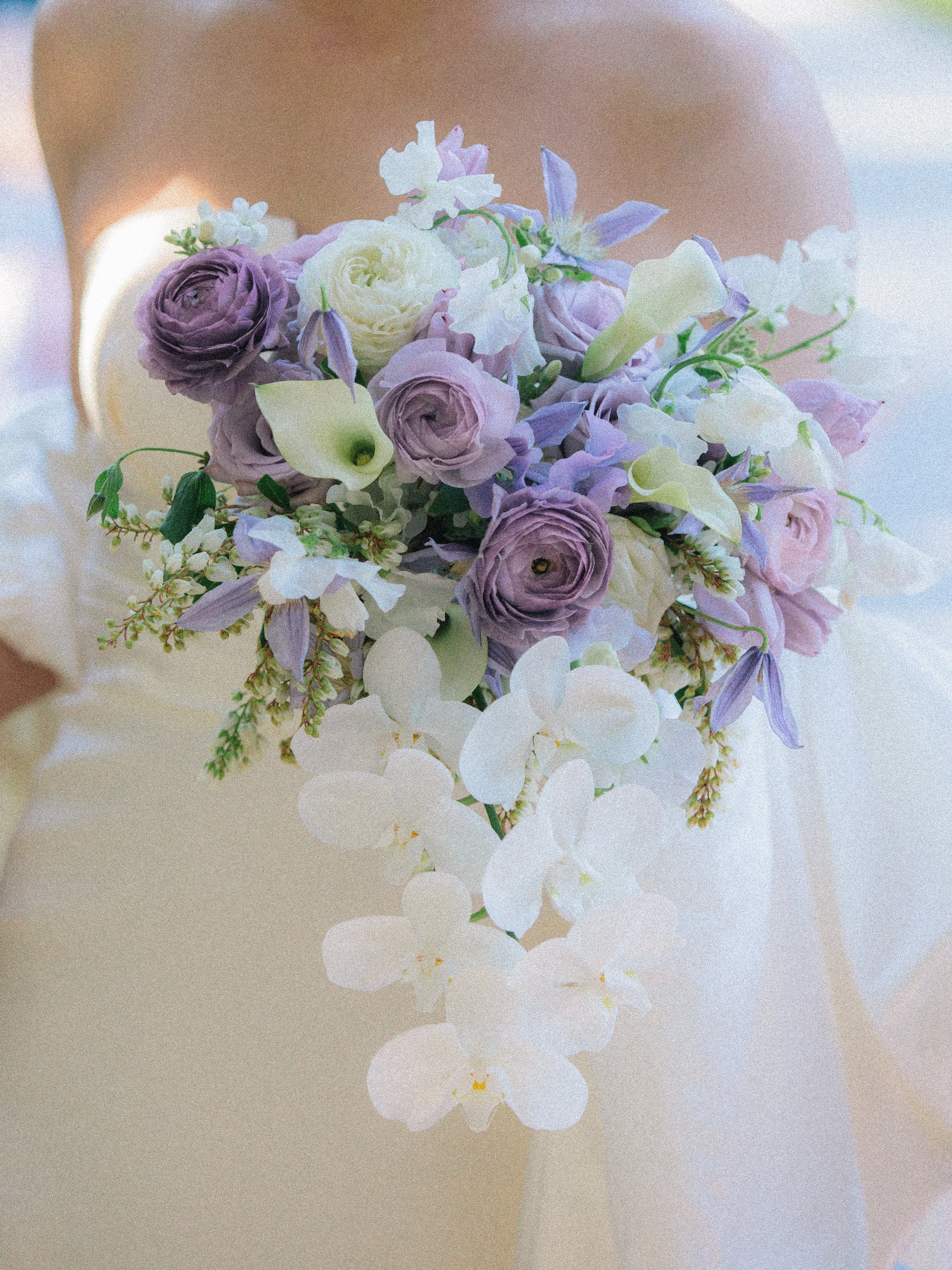 A bouquet of purple, white, and light purple flowers with large white and purple petals, and greenery, held against a light background.