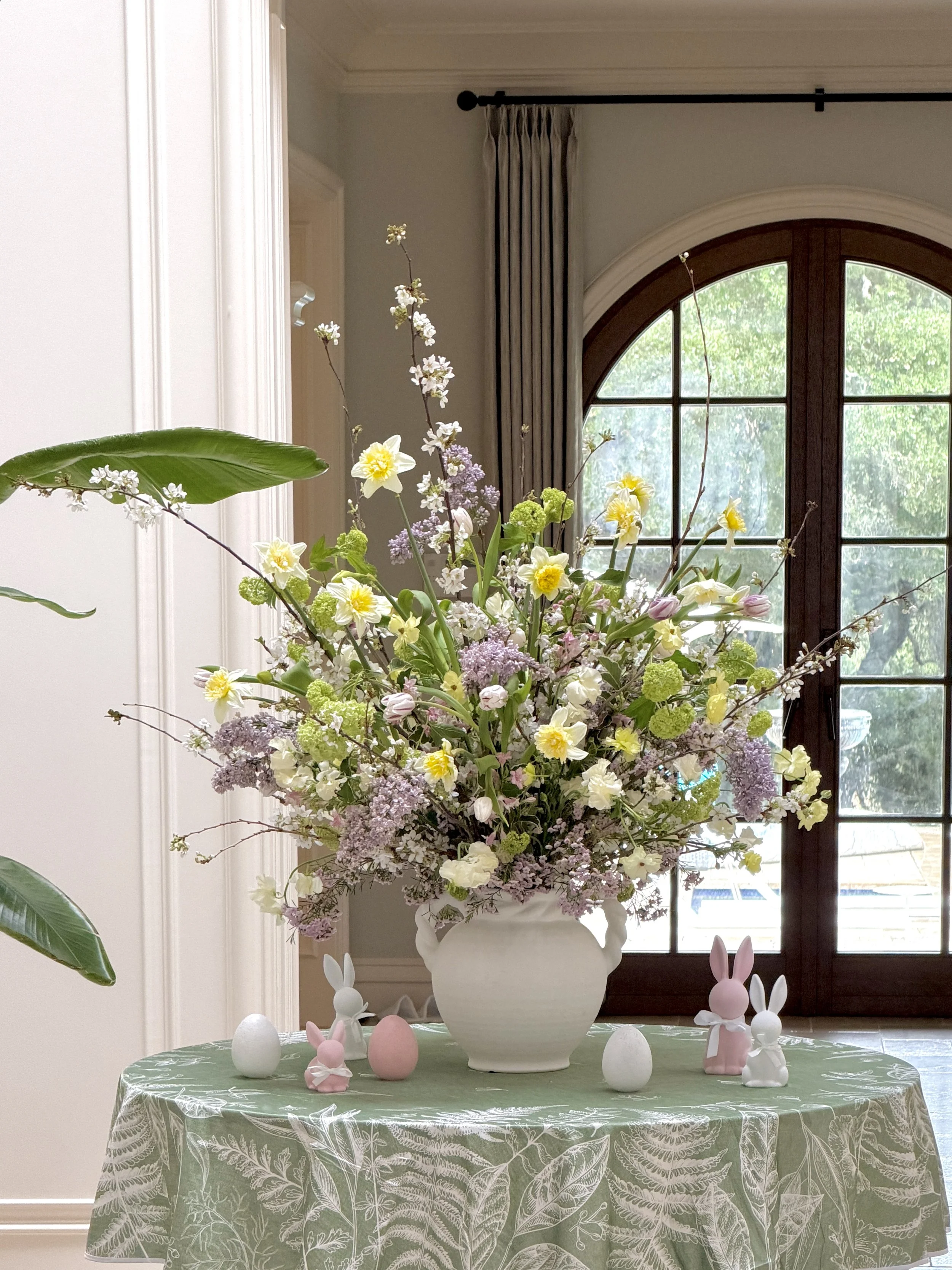 A large floral arrangement in a white vase on a table covered with a green tablecloth that has a white leaf pattern. Decorations resembling pink and white bunnies and eggs are placed on the table, which is in front of a window with a curved top, wood