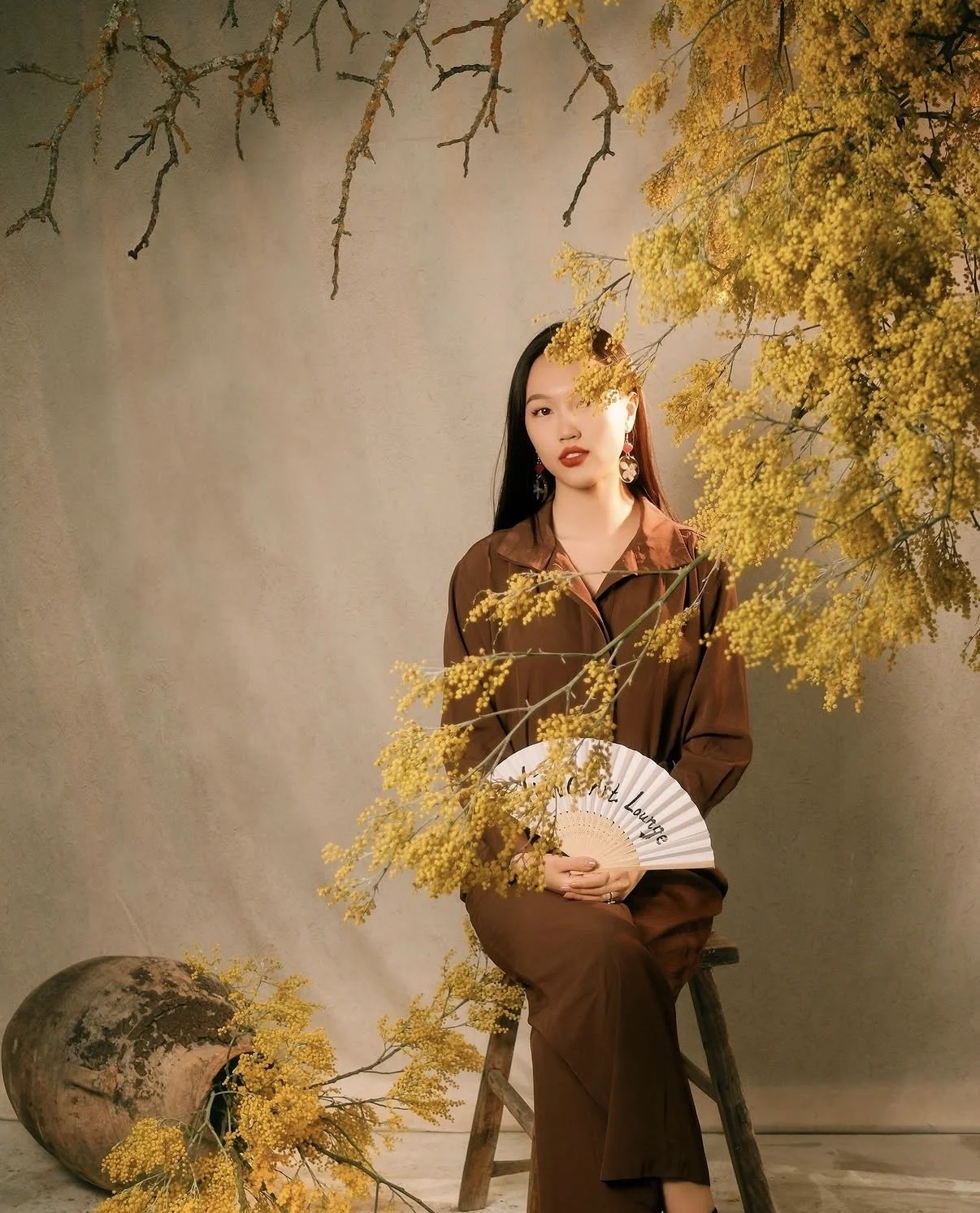 A woman sitting on a wooden ladder holding a paper fan surrounded by yellow flowers and branches, with a plain beige background.