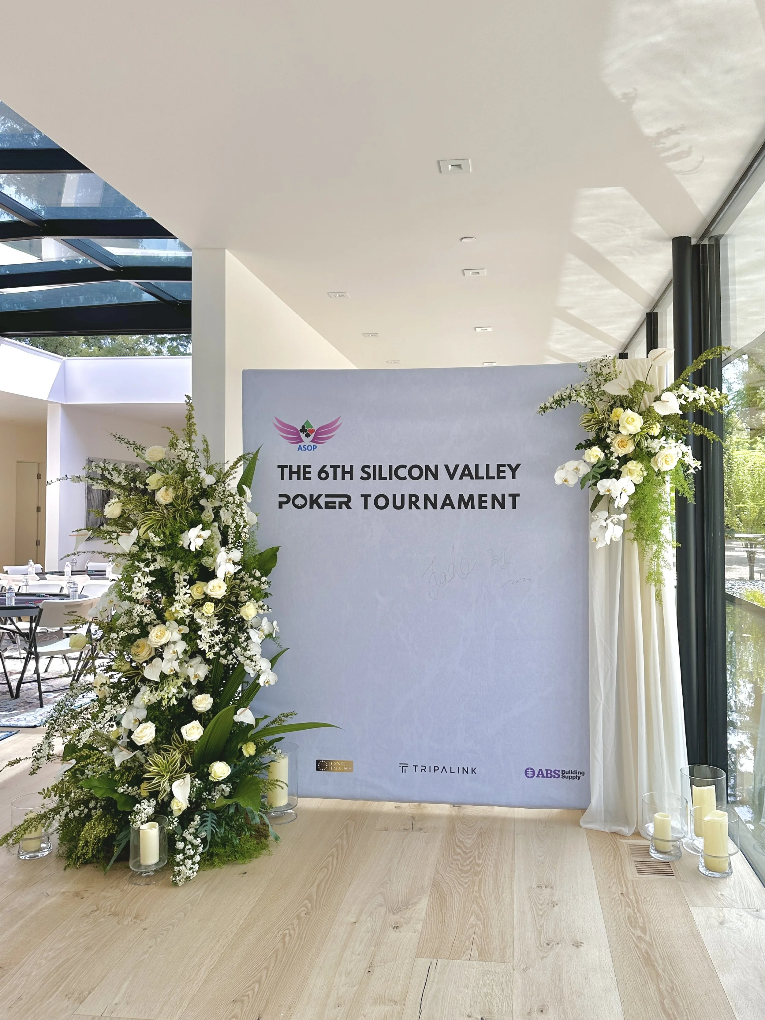 Decorative floral arrangements with white roses and greenery beside a sign for the 6th Silicon Valley Poker Tournament, with logos of Triple Link and ABS Building Supply at the bottom.