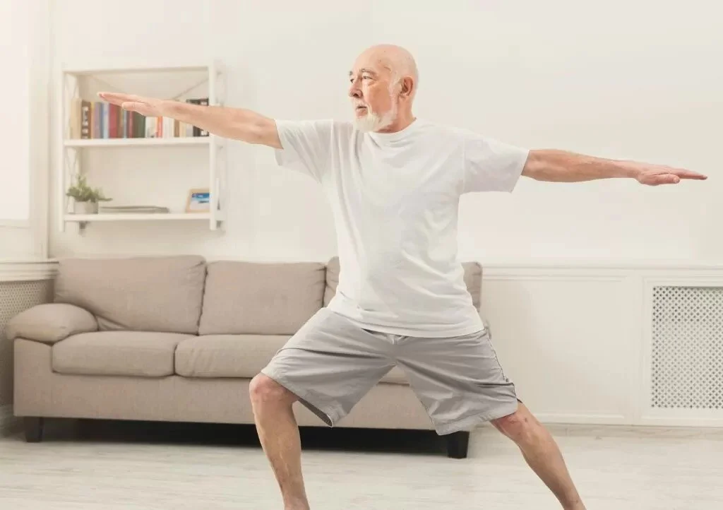 An elderly man in white t-shirt and shorts doing yoga stretches in living room.