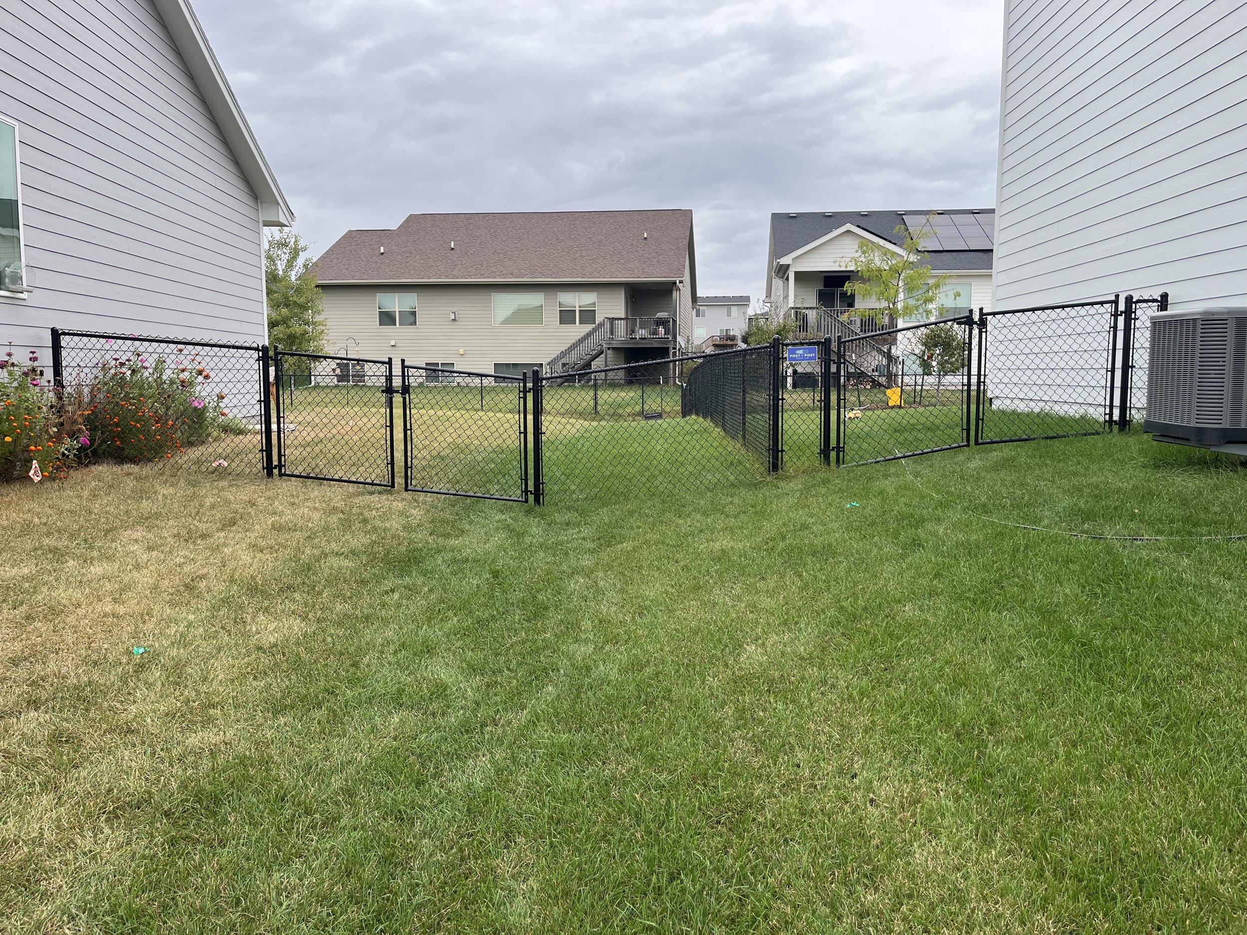 Two 4-foot black chain link fences installed on neighboring properties in Grimes, Iowa, tied together for a seamless connection. Both fences follow the sloped grade precisely, including properly aligned gates for a clean, continuous look.
