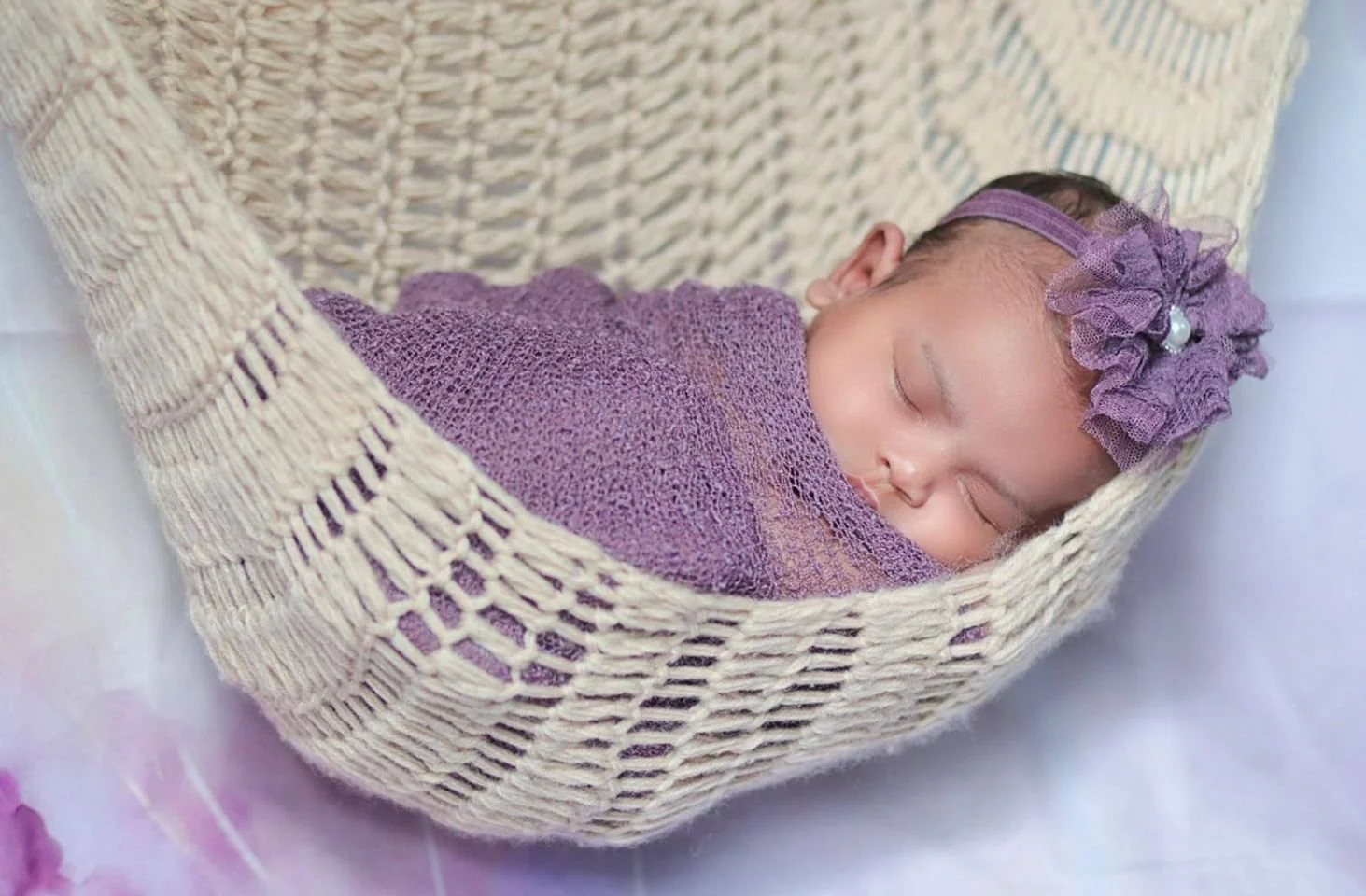 A sleeping newborn baby wrapped in a purple blanket, wearing a purple headband with an ruffled flower and pearl detail, resting in a cream-colored, woven basket.